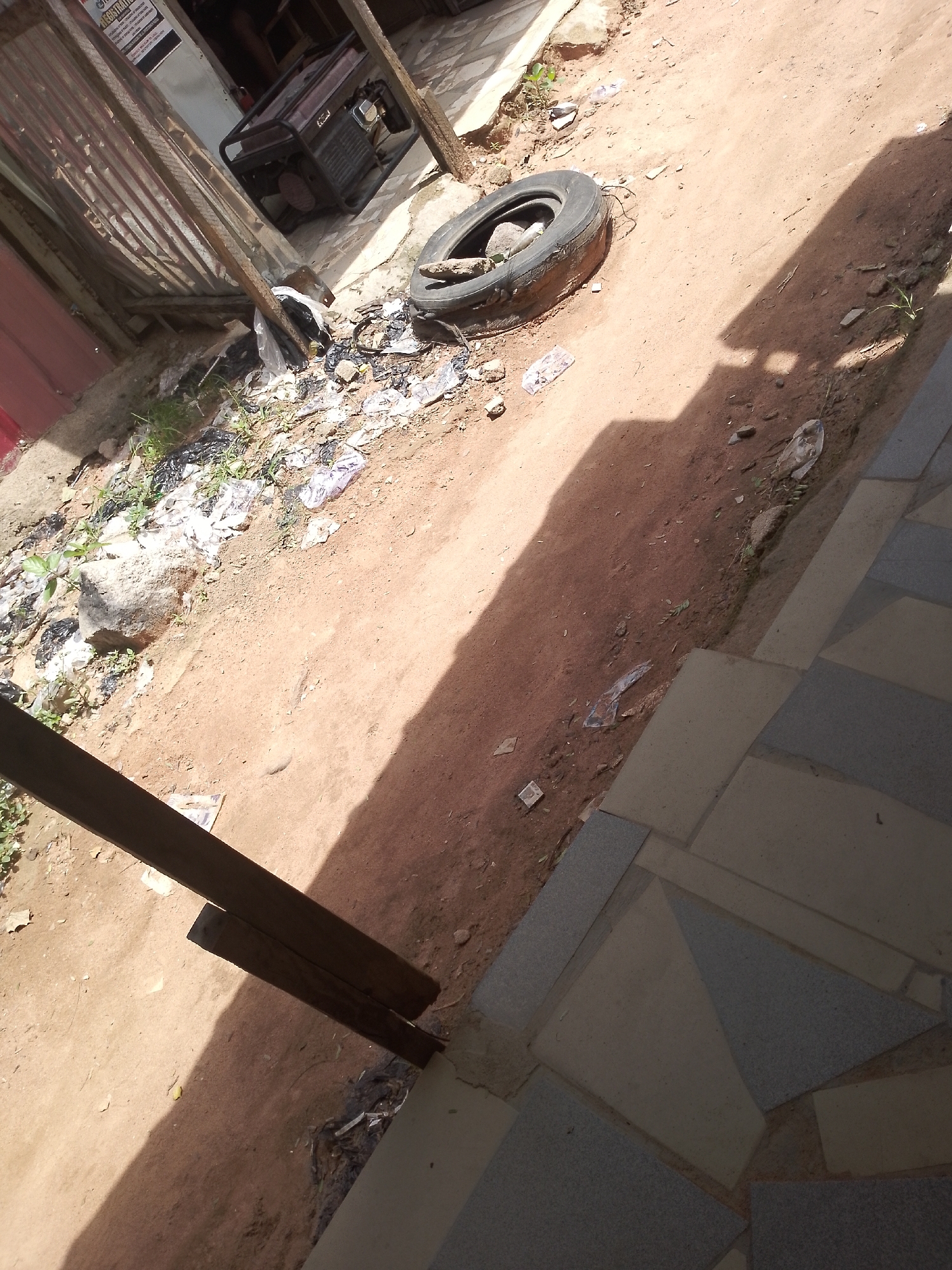 The image depicts an outdoor scene with a dirt ground, littered with various debris including plastic bottles, plastic bags, and what appears to be a discarded tire. In the background, a generator is visible, situated near a corrugated metal structure. A shadow, cast by an unseen object or structure, dominates the right side of the frame, creating a stark contrast with the sunlit areas. The foreground shows a paved walkway with irregularly shaped tiles. The overall impression is one of an unkempt urban environment.
