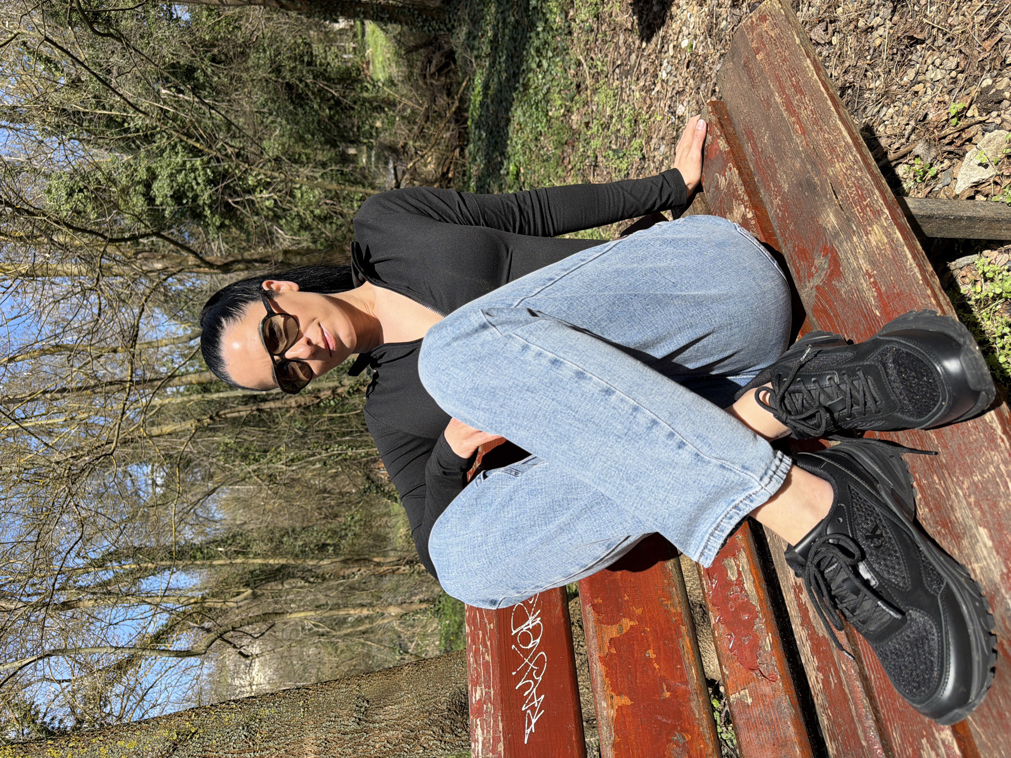 The image depicts a female subject seated on a weathered wooden park bench within a wooded area, specifically located in Pleven, Bulgaria. The subject, with dark hair pulled into a ponytail and wearing dark sunglasses, is dressed in a black long-sleeved top with a partially unzipped collar, light blue denim jeans, and black lace-up athletic shoes. She is positioned on the left side of the bench, with her legs crossed and her left hand resting on the bench behind her. Her gaze is directed forward. The wooden bench exhibits significant wear, with chipped and faded red paint revealing the natural wood underneath; white spray-paint graffiti, possibly reading "PAUFOR," is visible on the backrest. The background consists of a dense arrangement of deciduous trees, largely bare of foliage, with some green undergrowth and a distinct blue sky visible through the canopy. Strong natural sunlight illuminates the scene, casting shadows. The ground around the bench is covered with dry leaves, twigs, and sparse green vegetation. A large tree trunk with textured bark and visible lichen is present on the far left of the frame.