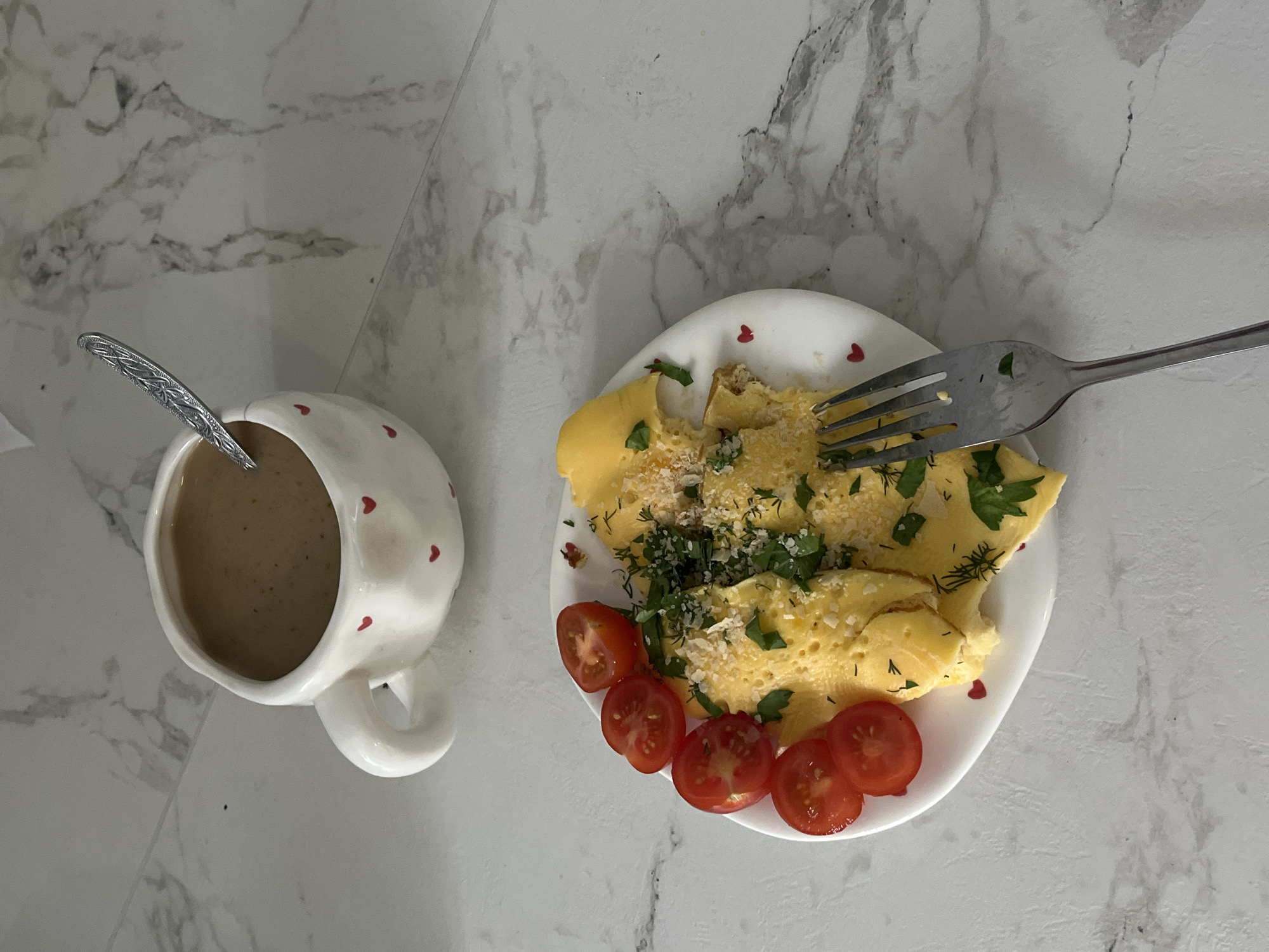 The image presents a breakfast scene, likely indoors. A white mug with red heart decorations, filled with a light brown liquid (possibly coffee or a latte) and a spoon inside, sits next to a small white plate. The plate holds an omelet, sprinkled with herbs and parmesan cheese, accompanied by sliced cherry tomatoes. A fork rests on the omelet. The setting appears to be a light-colored marble or tiled surface. There is no information to confirm that the location is Chaikovsky, Russia.