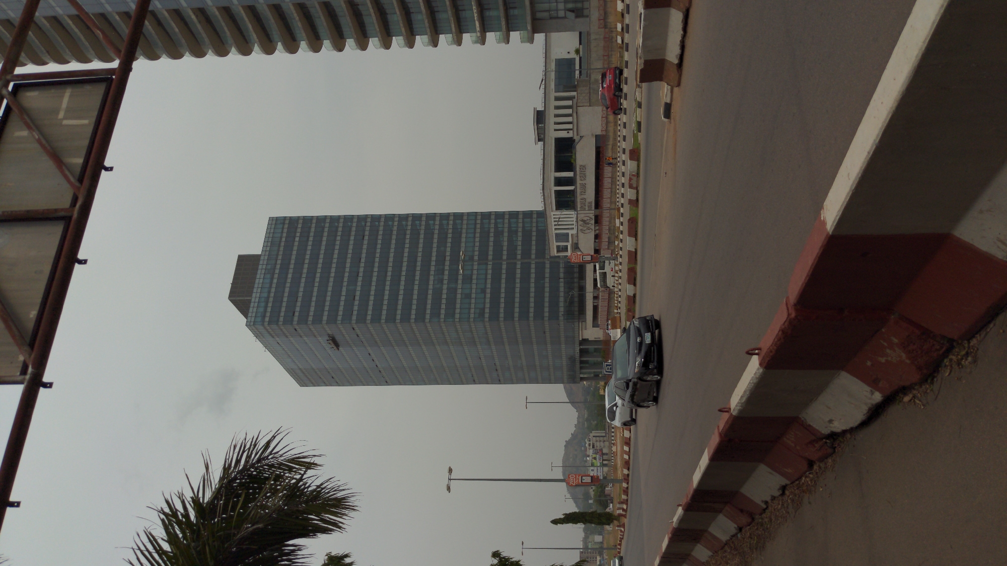 The image displays an urban landscape, likely captured on an overcast day, which can be identified as Abuja, Nigeria, based on visible text. The main subject is a tall, modern skyscraper dominating the left side of the frame. This building features a facade of blue-tinted glass panels arranged in a grid pattern, giving it a sleek, contemporary appearance.
To the right of the skyscraper, a multi-lane road stretches into the distance, with several cars visible, including a black car and a red car. The road is bordered by a series of concrete traffic barriers, distinctly painted with alternating red and white stripes.
Further to the right, behind the barriers, stands a lower-rise commercial building. This building prominently displays the text "WORLD TRADE CENTER" and "ABUJA" on its facade, confirming the location. In the background, beyond the urban development, distant hills are visible under the cloudy sky.
In the lower-left foreground, a large palm tree frond is visible, adding a touch of tropical vegetation to the urban setting. A metallic framework, possibly part of a large sign or overhead structure, is partially visible in the upper-left. Streetlights are also dotted along the road. The overcast sky suggests subdued lighting, and the general atmosphere is one of a bustling commercial district.