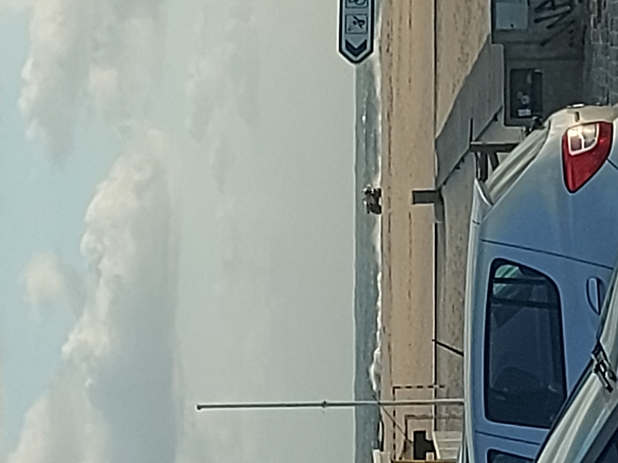 A sideways view captures a section of Figueira da Foz, Portugal's shoreline. To the right, the rear of a silver hatchback car is visible, parked along a paved area next to a brick wall. Further to the left, the shoreline stretches into the distance, bordered by the ocean. Waves are visible at the water's edge. The sky is cloudy. A sign indicating beach access with a downward arrow is positioned above the beach. In the middle distance on the beach, two small, dark figures are discernible. A pole extends horizontally from the bottom center towards the left.