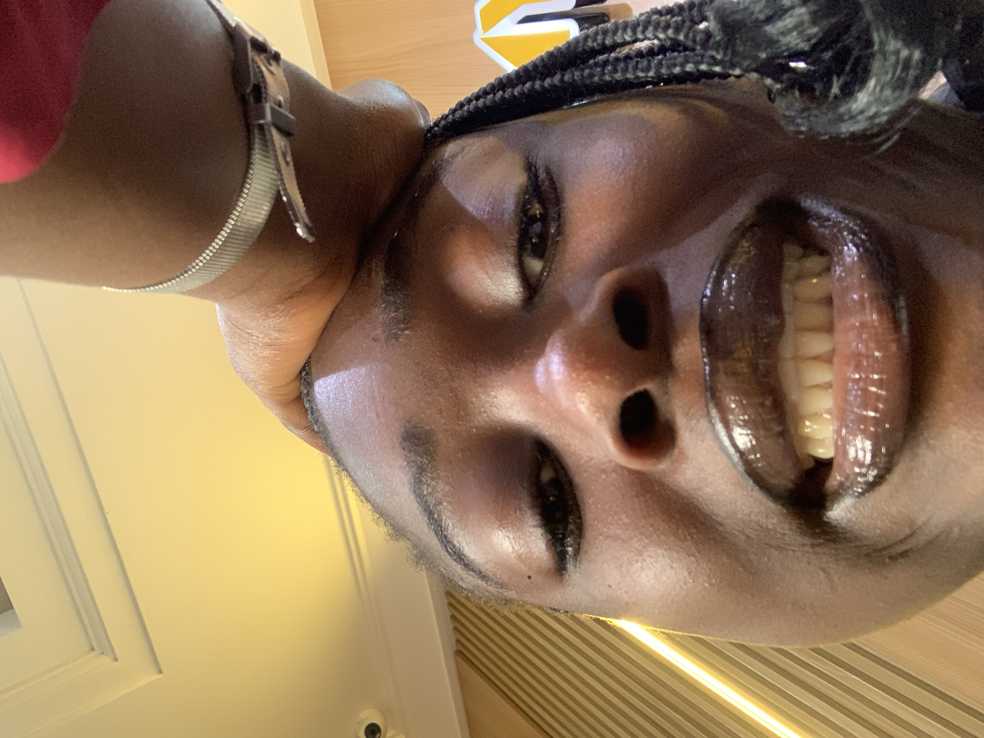 A close-up, low-angle shot captures a young Black person's face, looking upwards. Their mouth is slightly open, revealing teeth, and their lips are covered in a dark, glossy lipstick. A dark braid rests on their right shoulder, and a silver watch is visible on their left wrist. The person's forehead is touched by another hand. The background features a yellow ceiling with molding, a security camera, and a wall with vertical wooden panels. Yellow lights are visible on the left side of the frame. The scene is indoors.