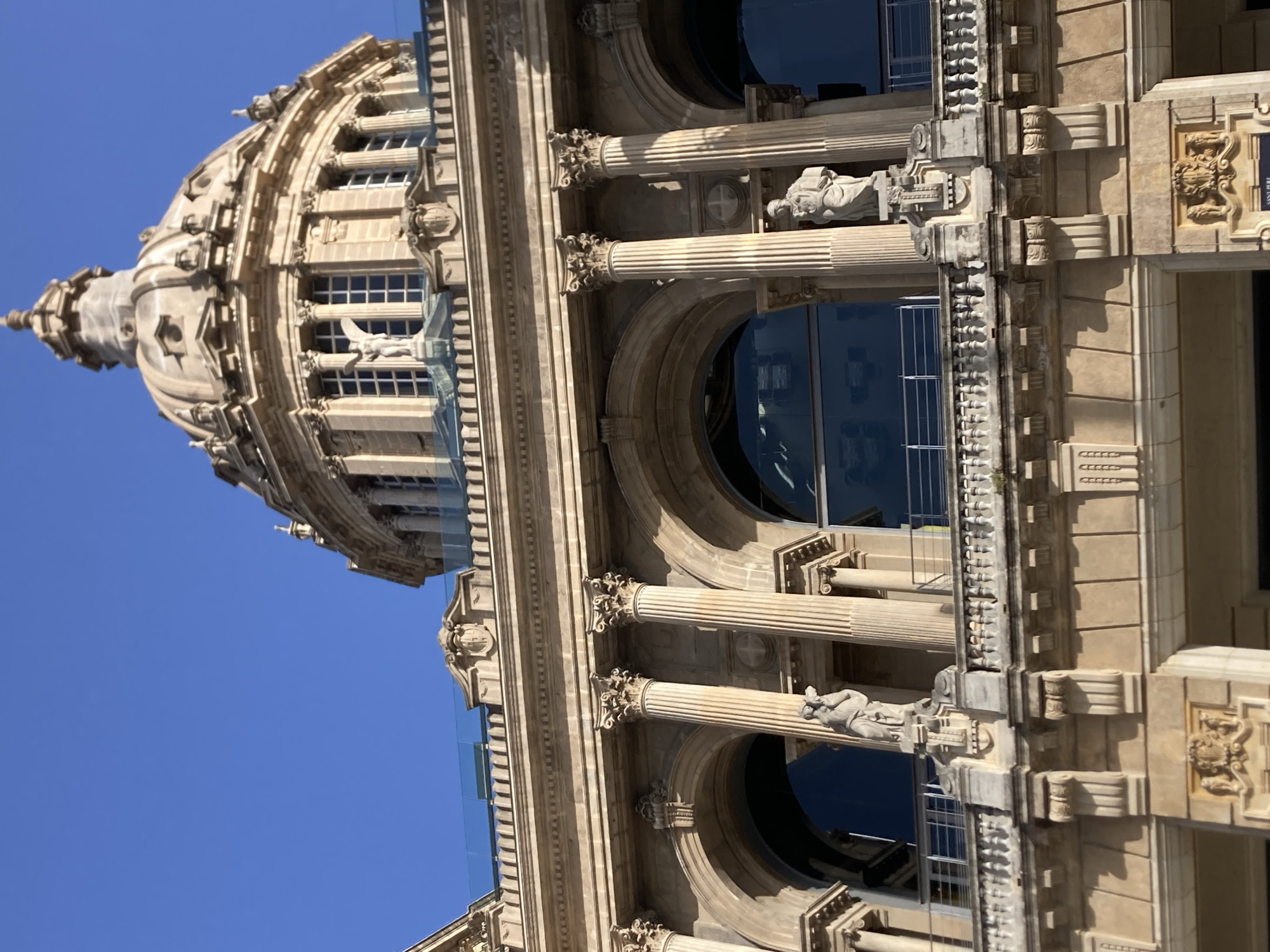 The image shows a close-up view of the National Palace of Montjuïc in Barcelona, Spain. The building features a grand dome at the top, adorned with architectural details. Below the dome, there are multiple levels of classical architecture, including columns, arches, and ornate sculptures. Several statues of figures in classical attire are visible along the balconies and alcoves of the building. The facade is made of light-colored stone, and the sky behind is a clear, bright blue. The architectural style suggests a historical and monumental building, consistent with the National Palace, which houses the National Art Museum of Catalonia.