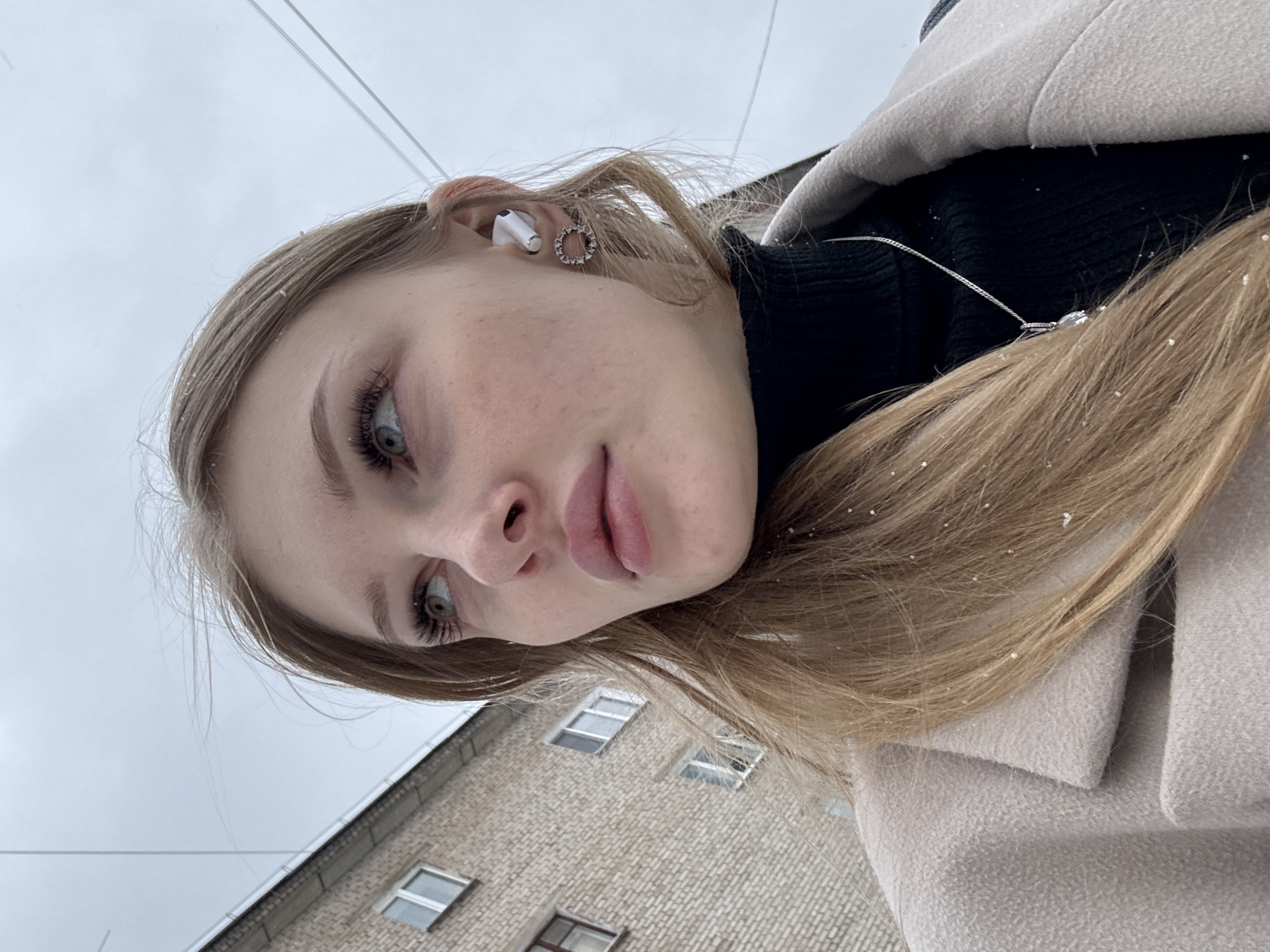 A young woman with blonde hair is looking into the camera at an upward angle. She has an earbud in her left ear and is wearing a black turtleneck, a light beige coat, and a silver necklace. Snowflakes are visible on her hair and clothing. The background includes a brick building with windows and a cloudy sky. The location is likely Perm, Russia, based on the architectural style of the building, clothing and likely weather conditions.
