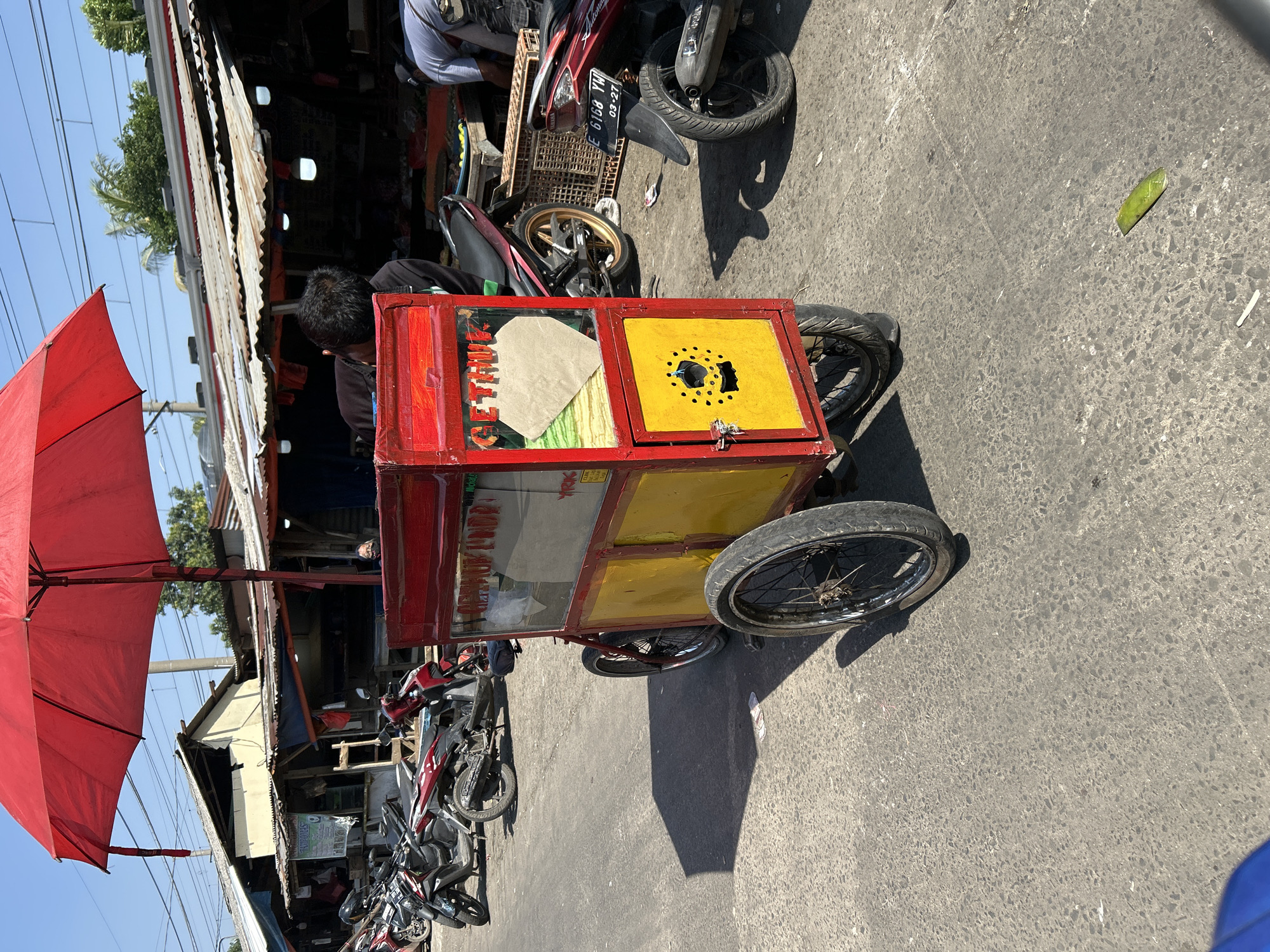 This is a medium shot, eye-level, outdoor, daytime photograph taken on a street in Depok, Indonesia. A red and yellow pushcart with an umbrella is positioned centrally in the frame. The cart has two wheels visible, with a third wheel likely at the front, suggesting it's a tricycle-style cart. A partially obscured person in a black shirt is leaning over the cart, possibly interacting with its contents. Another person wearing a light-colored shirt is visible further back, behind the cart, near a market stall.
The market area appears to be a street-side setup with various stalls and corrugated metal roofs. Several motorcycles are parked in the background, indicating a typical urban street scene. The ground is paved asphalt, showing some wear and tear. A large red umbrella is open above the cart, providing shade. The cart itself has glass panels displaying goods, and a section with a yellow panel featuring a black, circular design with cutouts. The overall atmosphere suggests a bustling marketplace.