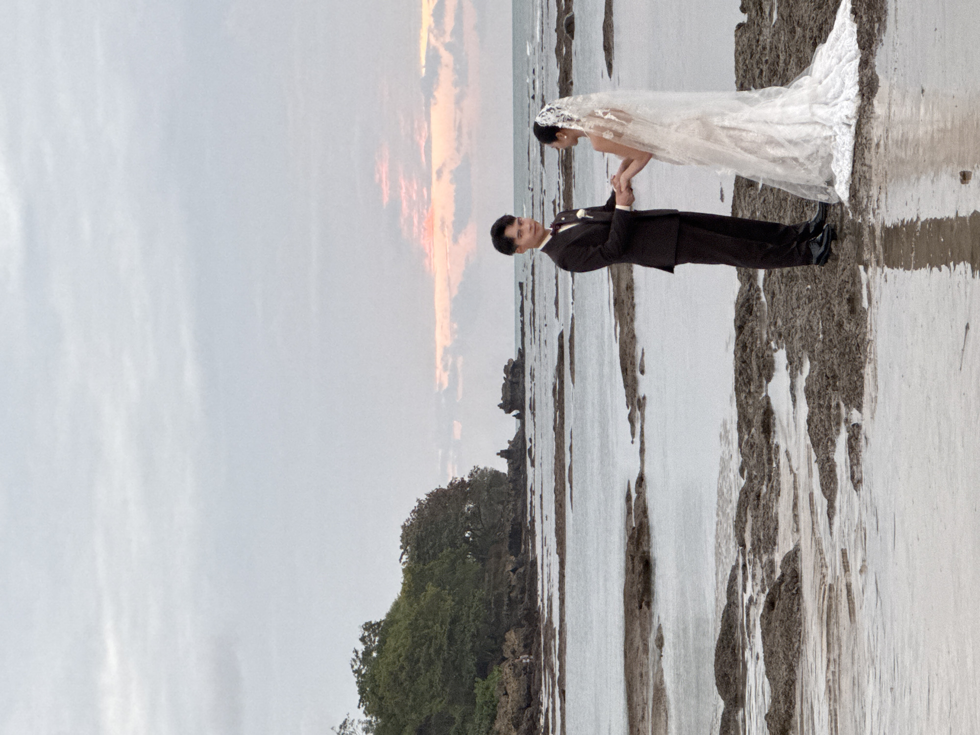A wedding couple, a man and a woman, stand on a rocky shoreline near Kuta Selatan, Indonesia, holding hands. The man is dressed in a dark suit with a bow tie and a white shirt, while the woman wears a white wedding dress and veil. The ceremony appears to be taking place during dusk, with soft, pastel colors painting the sky and clouds. The ocean is calm, with the tide out, revealing a rocky beach with tidal pools. In the background, a lush green treeline and rocky outcrops complete the natural, serene setting.