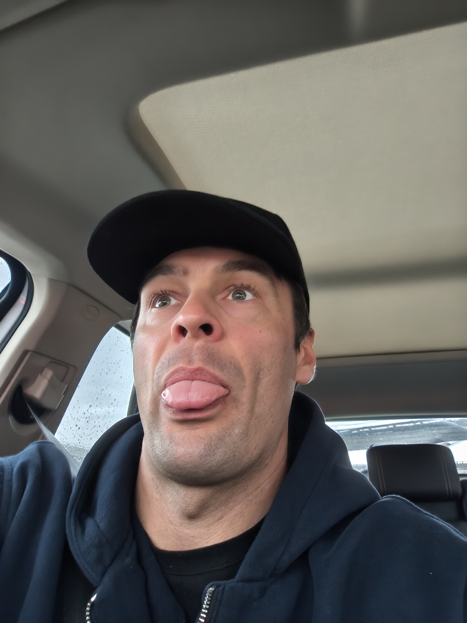 A man wearing a black cap and a navy blue hooded sweatshirt is inside a car, sticking his tongue out. The car appears to have a sunroof. Raindrops are visible on the car window. The location is not Moscow, Russia. There is no indication of the city or country.