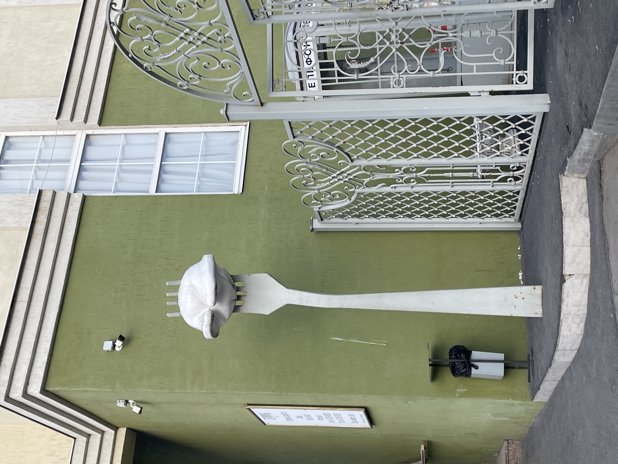 The image depicts a street-level corner of a building in Izhevsk, Russia, under ambient daylight conditions. The primary visible wall is painted light olive green, with a beige upper section featuring horizontal architectural moldings. On the green wall, two white surveillance cameras are mounted near the corner, and a rectangular white sign with Cyrillic text is affixed vertically to the left.
A prominent, oversized sculpture of a light-grey fork piercing a large, rounded, dumpling-like object stands on a raised platform edged with white tiles at the building's corner. The dumpling-like object features several small protrusions on its upper surface. To the right of the sculpture, a decorative, light-grey wrought-iron gate, adorned with scrollwork and a diamond lattice pattern, is partially open. Behind this gate, a grey public telephone booth is visible, marked with "ТЕЛЕФОН" in Cyrillic script on its upper panel.
Two white-framed, multi-paned windows with white curtains are visible on the beige upper facade. A narrow, light-colored trash receptacle containing a black plastic bag is mounted on a dark metal stand against the green wall, to the left of the sculpture. The ground surface is dark grey pavement, with a set of steps visible on the far left. No individuals are present in the scene.