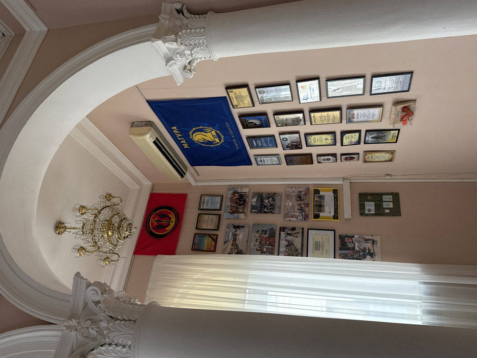 An interior scene depicts a room within a facility of the Magura martial arts and patriotic education organization in Kyiv, Ukraine. The room features a pale peach-colored wall with white architectural detailing, including two fluted columns with ornate Corinthian capitals and a wide, white arched ceiling element. A multi-tiered crystal chandelier with gold-toned metal components hangs from the white ceiling centrally within the archway.
On the left side, a large window is covered by sheer white curtains, providing diffused light. Adjacent to the window, a red flag with a black circular emblem depicting a gold winged sword and Cyrillic text reading "ЮНОША МАГУРА" (Youth Magura) and "ОРЛИЙ ЛЕГІОН" (Eagle Legion) is displayed. Below this flag, a collection of framed photographs, primarily showing groups of young individuals, and framed certificates, including one marked "ГРАМОТА" (Diploma/Certificate), are arranged on the wall. A small olive-green display case containing multiple badges or pins is mounted lower on this section of the wall.
On the right wall, an off-white wall-mounted air conditioning unit is installed high. Beneath it, a blue flag prominently displays a yellow emblem of a stylized knight chess piece with the number "47", along with Cyrillic text "МАГУРА" and the Latin phrase "SEMPER AUDENTES!" (Always Daring!). This wall is extensively covered with a grid of various framed certificates, awards, and plaques of differing sizes and frame styles, many bearing Cyrillic inscriptions. Below these, a small clear plastic box containing red ribbons or decorative items is affixed to the wall. No individuals are present in the immediate scene.