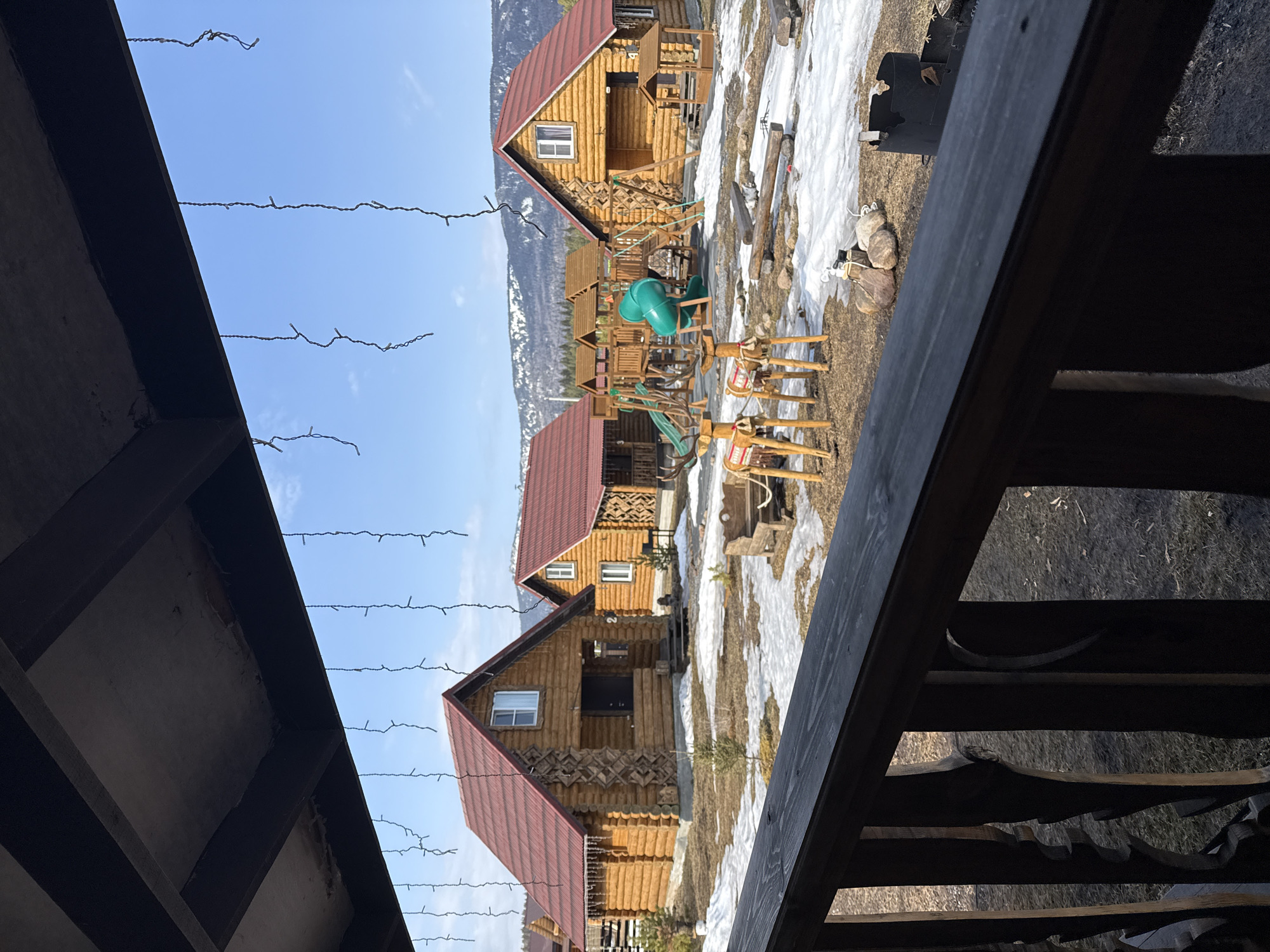 The image presents an outdoor scene in Tyulyuk, Russia, captured from a covered wooden porch or balcony. The viewpoint looks out over a dark wooden railing. Above, multiple strings of unlit, icicle-shaped decorative lights hang from a dark wooden ceiling structure.
In the midground, several log cabins with reddish-brown corrugated metal roofs are visible. The closest cabin to the left features a visible house number "2" near its dark entrance door, and a few icicles are present along its roofline. The ground is a mix of brown earth and melting snow patches. Small, bare shrubbery is scattered throughout the scene.
A children's playground structure, constructed primarily of wood with a prominent green tube slide, is situated between the cabins. Two decorative wooden reindeer figures, each adorned with patterned fabric saddles, stand facing right in the central midground. Other outdoor features include a rustic wooden bench, a stone-lined fire pit, and a black metal fire pit or brazier with a grate.
The background reveals forested mountains, some with visible snow-capped peaks, under a clear blue sky with minimal cloud cover. No individuals are present in the scene.
