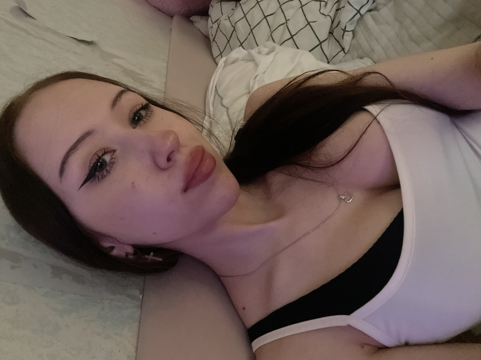 A young woman with long, dark hair lies on a bed, looking at the camera. She is wearing a white tank top over a black bra and a silver necklace with a heart pendant. She has on eyeliner and lip gloss. In the background, a textured wall is visible, along with a pillow with a checkered pattern. The room is dimly lit and appears to be a bedroom in Votkinsk, Russia.