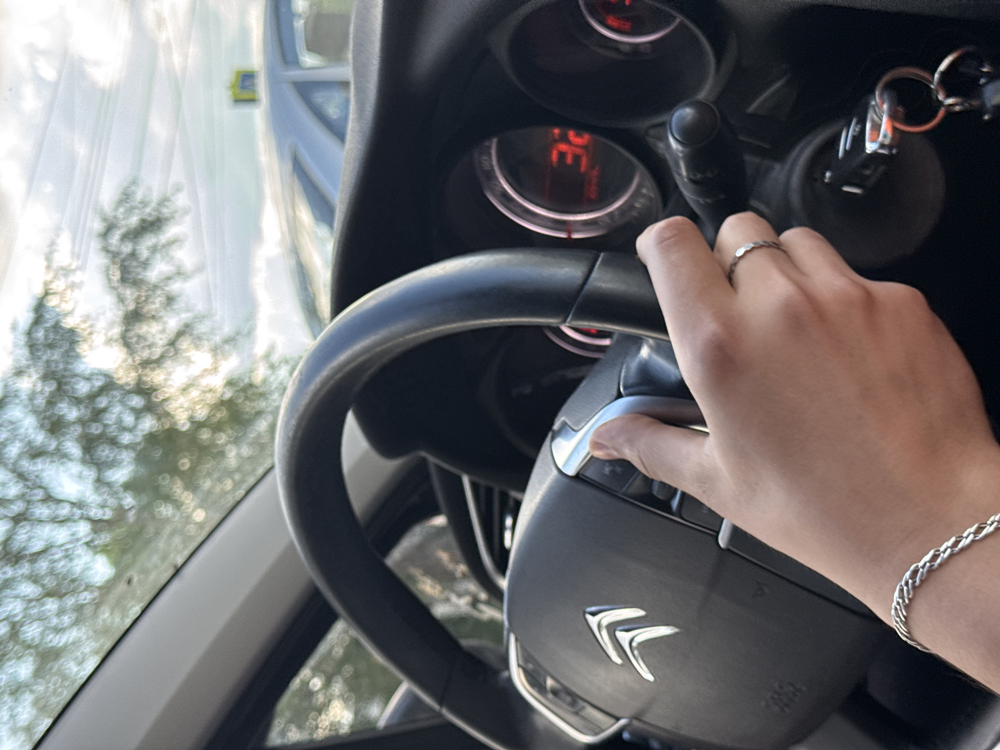 A medium-shot, high-angle photograph shows the interior of a Citroën vehicle, focusing on the steering wheel and the driver's hand. The steering wheel is black, with a silver Citroën logo prominently displayed. The driver's hand, adorned with a silver ring and a silver bracelet, is positioned on the steering wheel. The dashboard, with illuminated gauges showing "32", is visible in the background. Car keys are in the ignition. Through the windshield, blurred foliage and building are visible. There is no indication that this photo was taken in Izhevsk, Russia.