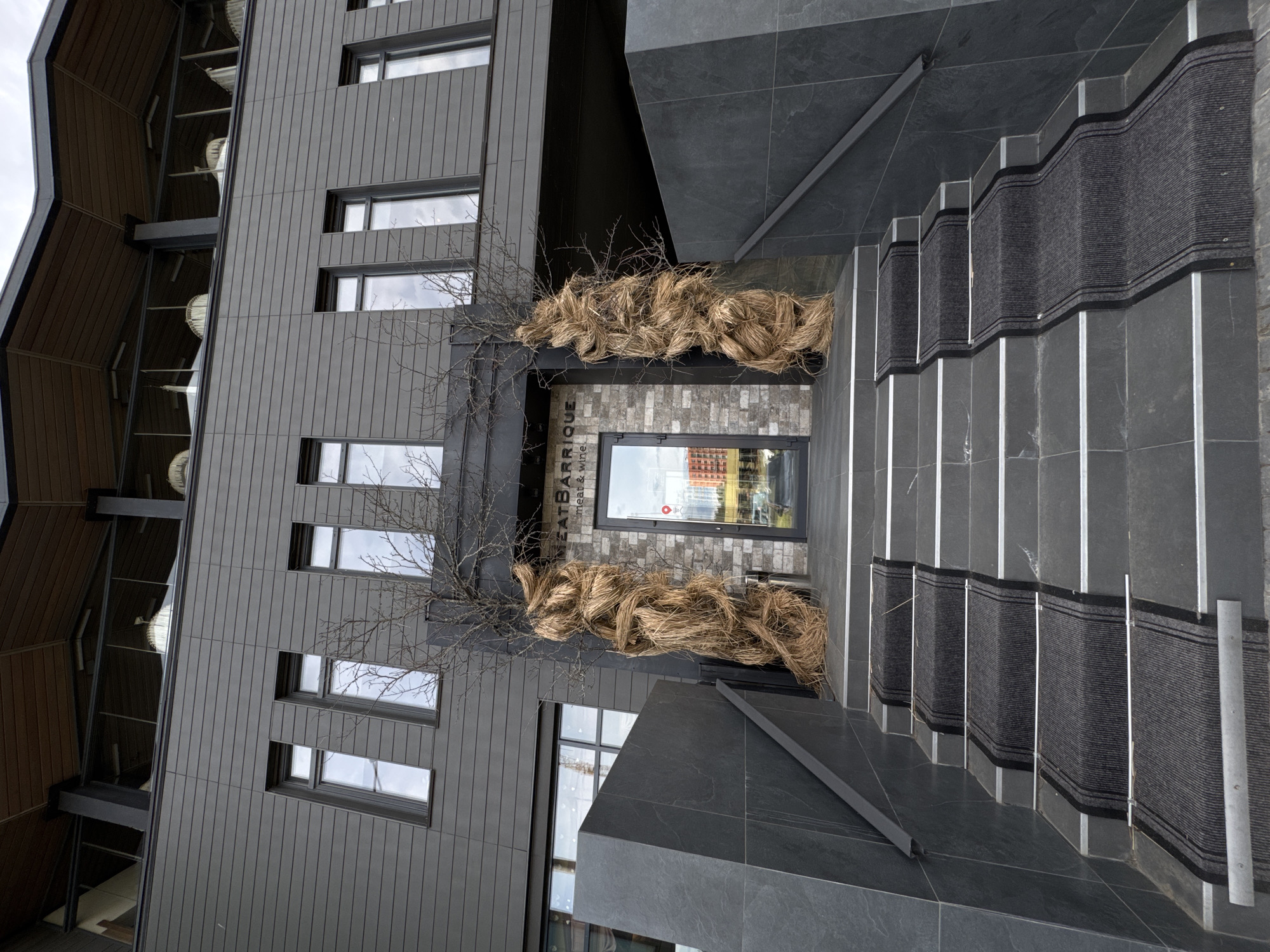 The image shows the exterior of a modern building in an unknown city. The building has a gray facade with rectangular windows. The entrance is decorated with dried branches and hay-like material. A set of stairs with dark gray tiles and carpeted risers leads to the entrance. The building appears to house "EAT.BARRIQUE meat & wine". The sky is overcast. The image is taken at an unusual angle, likely tilted, creating a slightly disorienting perspective.