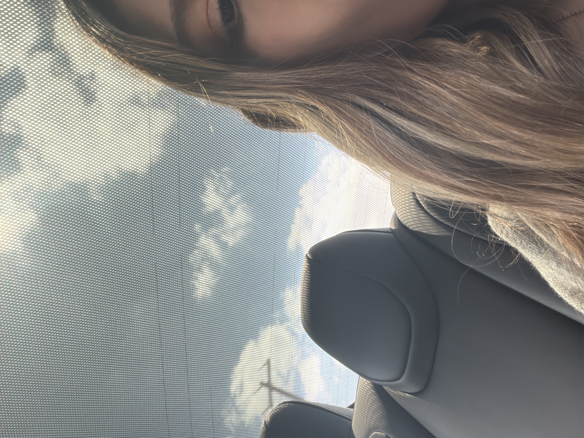 The image, seemingly taken from inside a vehicle in Austin, Texas, presents a close-up view of a person with light brown hair. The individual is partially visible, with only a portion of their face and hair in focus. The car's sunroof or window is visible, featuring a mesh or dotted pattern. Through the glass, a glimpse of the sky with some clouds is apparent. The interior of the vehicle includes a gray leather headrest of the car seat. An antenna is visible through the glass.