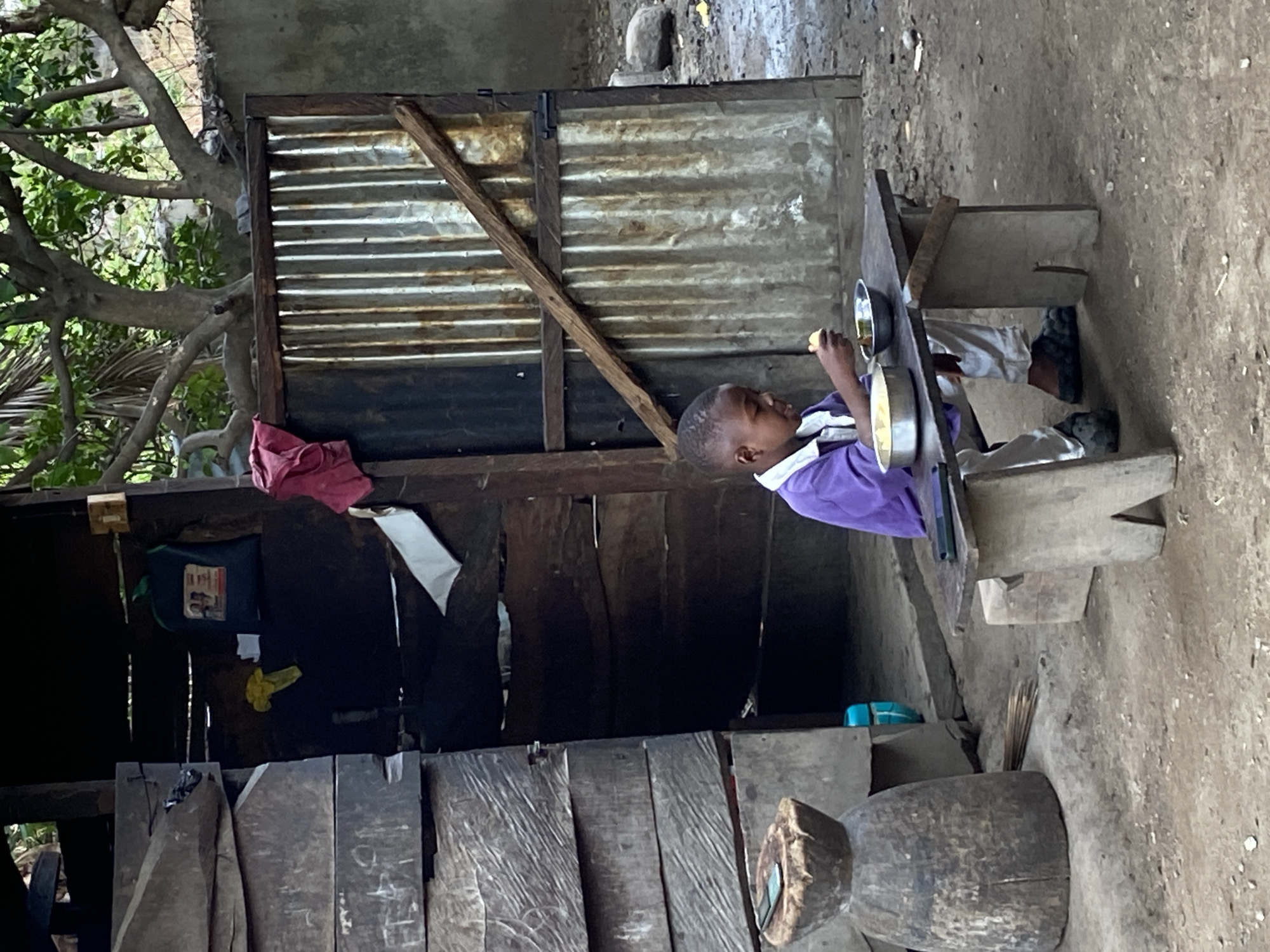 A young boy is sitting at a rudimentary wooden table eating from a plate in a likely rural or impoverished area. The boy is wearing a purple shirt and light-colored pants and shoes. The table is made of roughly hewn wooden planks, and he is seated on a wooden stool. Behind the boy is a makeshift structure constructed from wooden planks and corrugated metal sheets. Inside the structure, items are hanging, including what appears to be a bag and some clothing. The ground is dirt. While Mararaba, Nigeria, is a city, the surrounding areas can have these types of rural scenes.