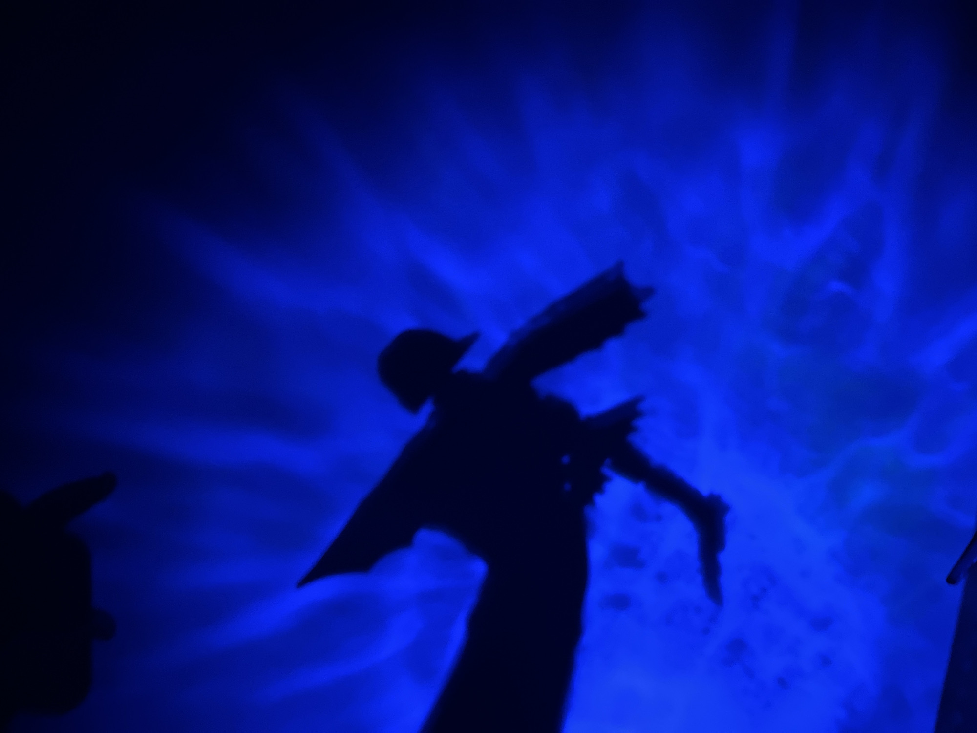 A silhouette of a person wearing a hat and jacket is projected onto a dark blue, illuminated surface, possibly a wall or curtain. The silhouette is in a dynamic pose, with arms outstretched, suggesting movement or dancing. The illumination creates a smoky, atmospheric effect, with streaks and wisps of light radiating outwards. The scene is dark overall, with the bright blue light being the primary source of illumination. There is also a dark shadow of another figure or object in the upper left corner of the image. The overall impression is one of artistic expression or performance, possibly in a nightclub or theater setting. There is no information within the image to indicate a specific location in Saint Petersburg, Russia.