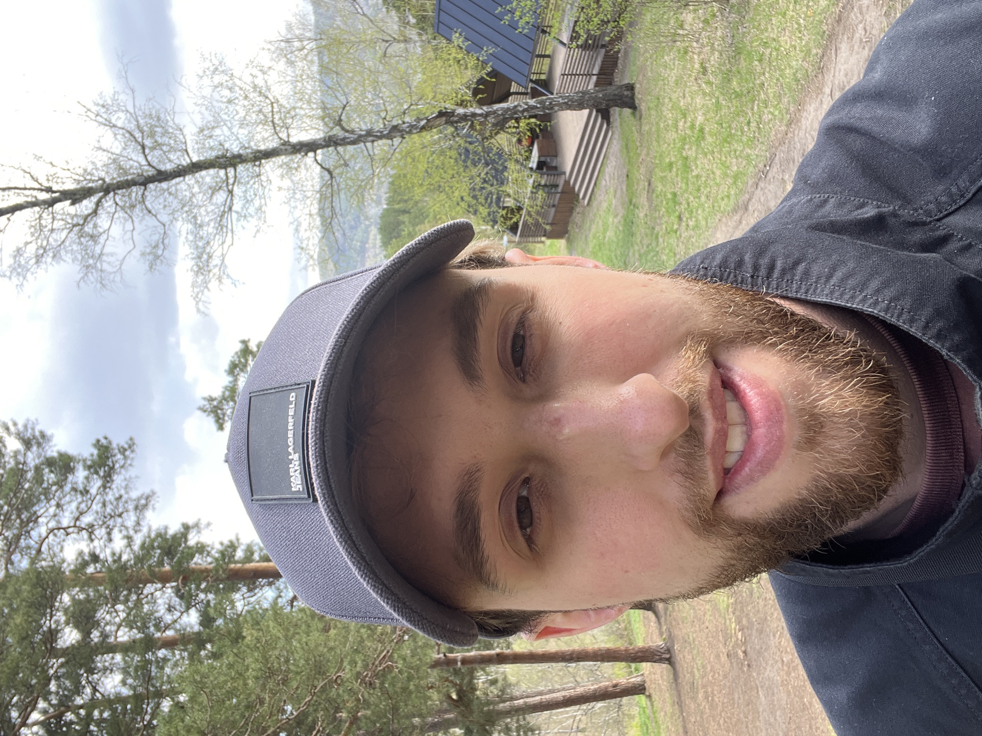 A person with fair skin, light brown hair and beard, wearing a grey Karl Lagerfeld baseball cap and dark jacket, is taking a selfie outdoors. The background includes trees, grass, and a building with a dark roof and stairs. The location appears to be a wooded area, potentially a park or recreational area. The specific location within Krasnoyarsk, Russia, cannot be determined solely from this image.