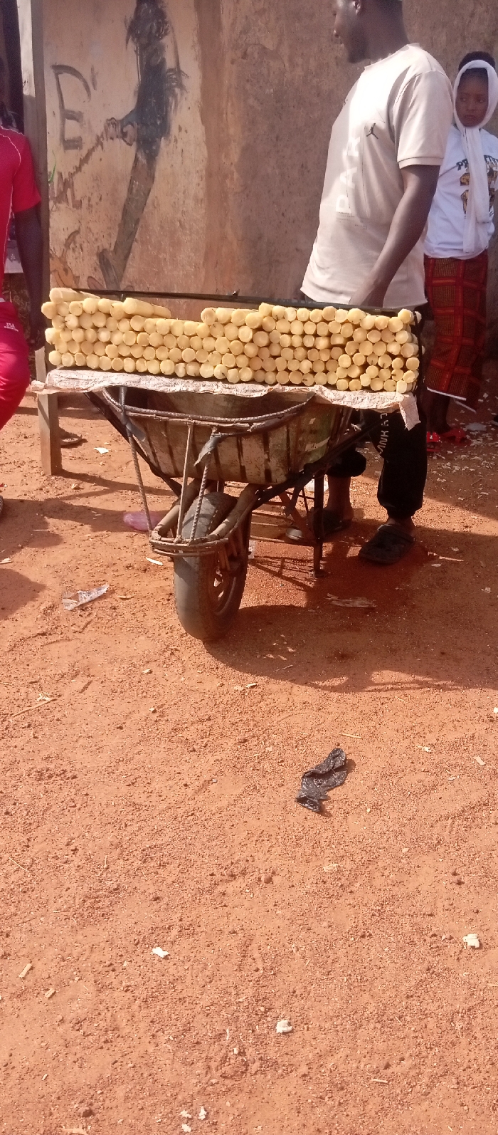 This image captures an outdoor street vending scene in the city of Kasuwan Magani, Nigeria. A metal wheelbarrow, appearing repurposed, supports a vendor's stand draped with a light-colored tarp. On this stand, numerous light yellow, cylindrical pieces of peeled cassava or similar root vegetable are neatly stacked in horizontal rows.
To the right of the stand, an adult male with dark skin stands facing left, wearing a light-colored t-shirt featuring a "Jumpman" logo and dark-colored trousers or track pants, along with black slide sandals. Behind him, a young girl with dark skin is partially visible, wearing a white head covering or hooded top and a patterned long skirt. On the far left, a portion of another individual wearing a red short-sleeved top is visible.
The ground is composed of reddish-brown dirt, with scattered small debris including white paper scraps and a crumpled black plastic bag. In the background, a light brown building wall, showing cracks and wear, features dark graffiti or a painting, including a large letter 'E' and a stylized dark figure. The scene is illuminated by bright natural sunlight, creating distinct shadows.