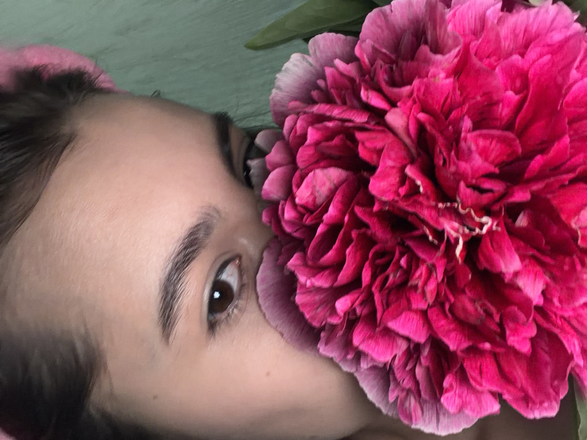 A close-up, horizontally oriented image shows a person's face partially obscured by a large, vibrant pink flower. The person has fair skin, a visible eye with brown iris and eyebrow. A petal of the flower appears to be resting on the person's nose. The background is a blurry green, which is consistent with foliage. Without additional information, I cannot confirm this image was taken in Chaikovsky, Russia.