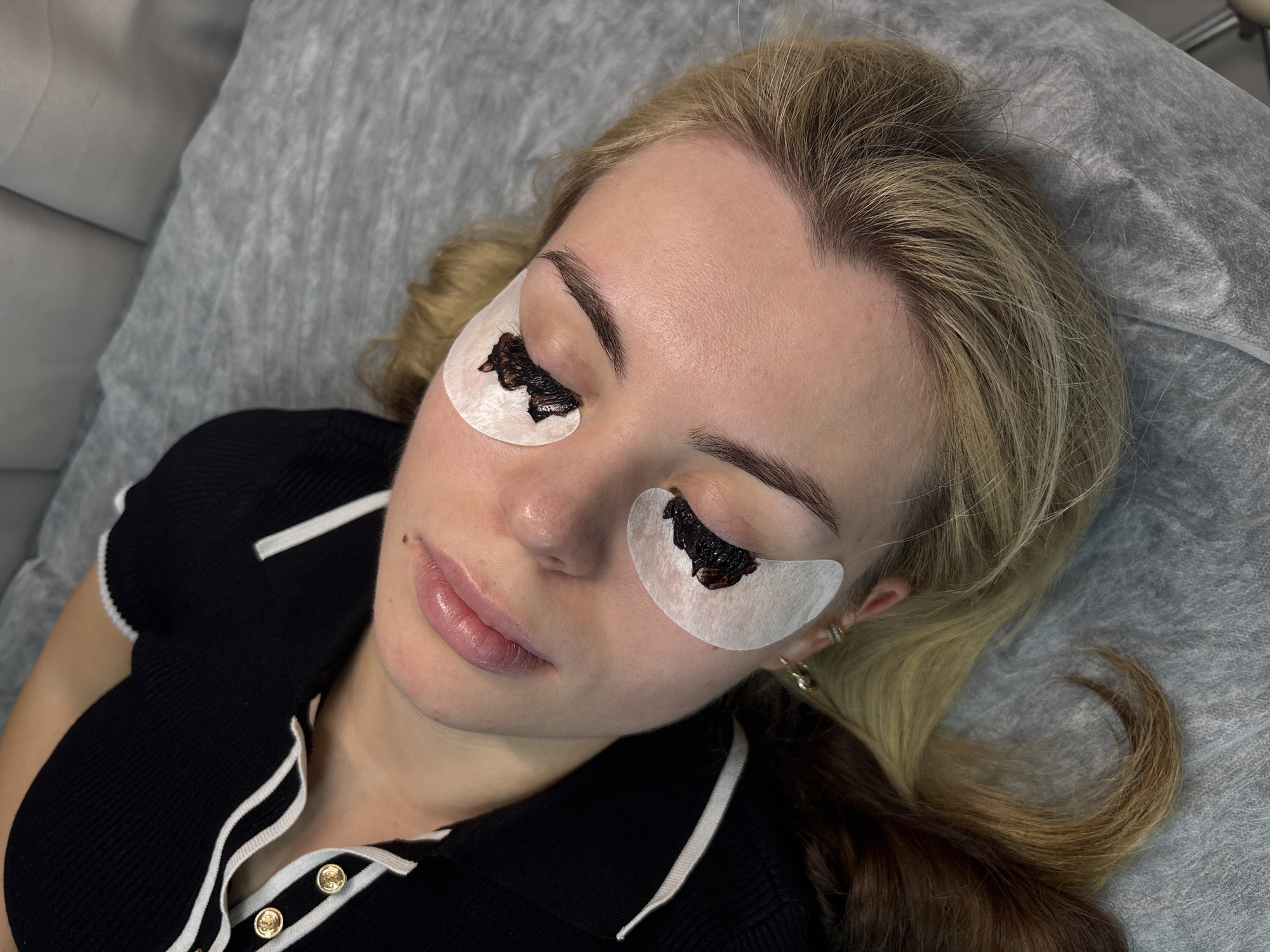 A young woman with blonde hair and fair skin is depicted from an overhead angle, undergoing an eyelash treatment in what appears to be a beauty salon. She is lying on a grey, textured, disposable cover, with her eyes closed. White, crescent-shaped under-eye pads are placed beneath her lower eyelids, on which a dark, paste-like substance—likely an eyelash tint or solution for a lash lift—is applied to her lashes.
The woman has defined eyebrows and glossy, natural-toned lips, with a small mole visible above the corner of her mouth. Her blonde hair is loosely styled, with some darker, possibly caramel or light brown, strands visible towards the bottom right of the frame. She is wearing a black, ribbed polo-style shirt detailed with white stripes on the collar and placket, featuring gold buttons. Small gold hoop earrings are visible in her ears.
The scene suggests a moment of professional beauty care, with the woman appearing relaxed during the treatment. The setting is indicative of an indoor beauty establishment, consistent with a salon environment in Perm, Russia. There is no visible text in the image.