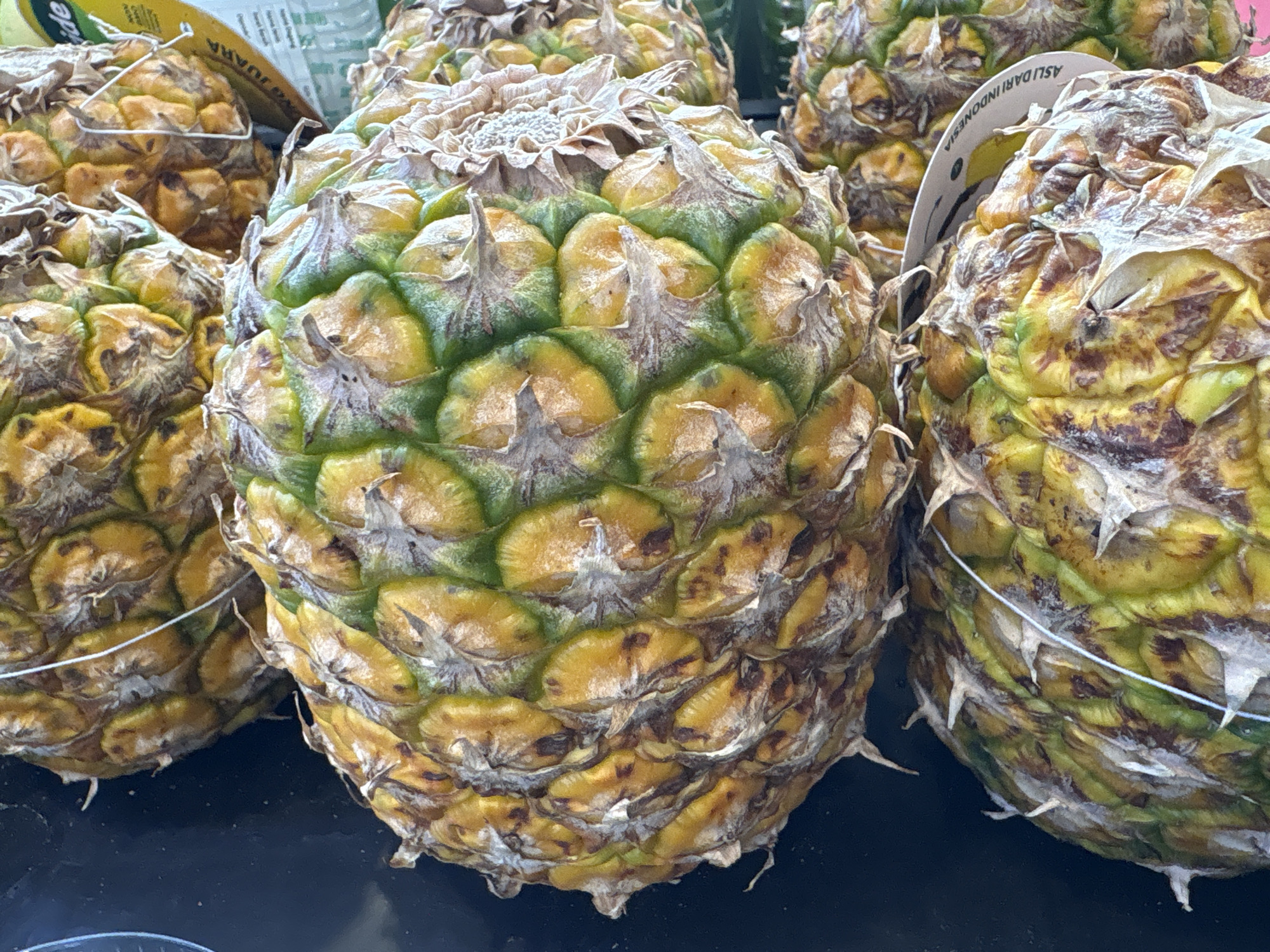 In a market setting in North Jakarta, Indonesia, several ripe pineapples are displayed. The foreground is dominated by the textured, spiky skin of the pineapples, which show a gradient of colors from deep green at the crown to yellow and brown on the body, indicating ripeness. Some of the pineapples have small white strings wrapped around them. In the background, partially visible, are other pineapples and parts of packaging or signage, one of which bears text that includes "ASLI DARI INDONESIA". The overall scene suggests a fresh produce display.