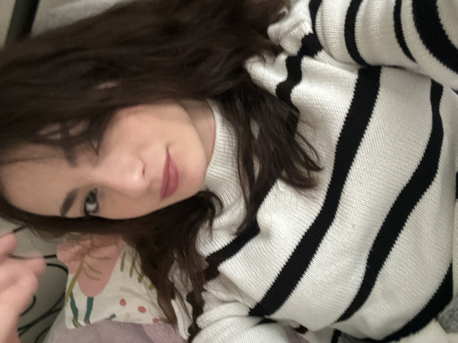 A close-up, chest-up portrait features a female individual with dark brown, wavy hair, dark eyebrows, and light-colored eyes, positioned against a soft, blurred indoor background. She is wearing a white knitted sweater with horizontal black stripes and a mock-neck collar. Her hair is parted on the left side, with strands falling across her left eye. Her lips are coated with a pink-mauve lipstick, and she exhibits a subtle smirk while looking directly forward. A blurred hand is visible at the upper left corner of the frame. A light-colored pillow with a faint floral pattern in pink and green hues is partially visible behind her head on the left. The environment appears to be an indoor domestic setting. This scene is located in Strasbourg, France.
