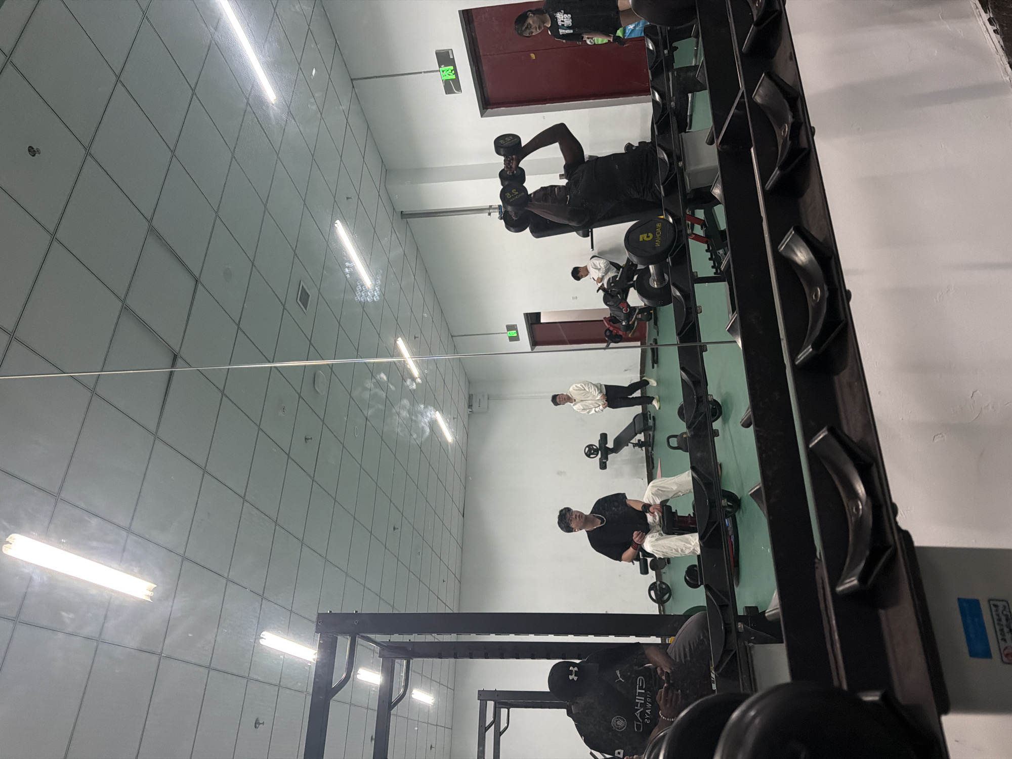 The image shows the interior of a gym. The location is inferred to be in Jiangning District, China. Several people are present, some engaged in weightlifting activities. One individual is seen lifting dumbbells, while others are using various exercise machines. A dumbbell rack is visible in the foreground. The gym features bright fluorescent lighting and mirrors along the walls. The scene includes exercise equipment such as benches, weight machines, and racks. The environment appears clean and well-maintained.