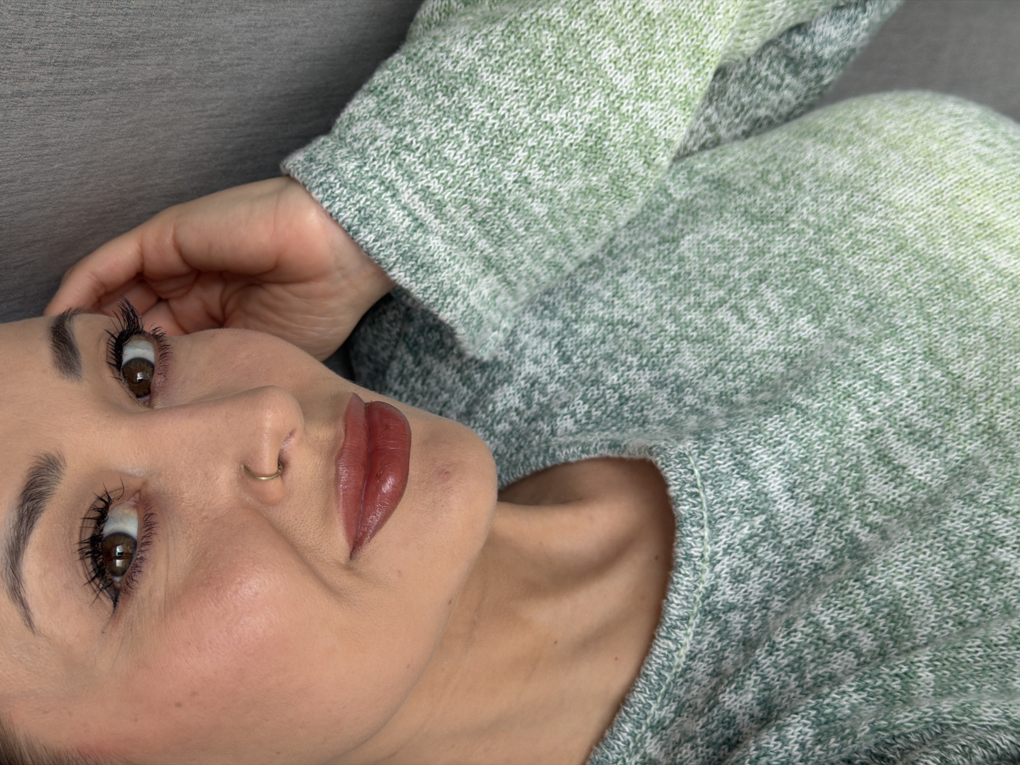 The image shows a woman lying down, likely on a soft surface like a couch or bed, wearing a green and white knit sweater. Her face is angled towards the viewer. She has brown eyes, long eyelashes with mascara, and a gold nose ring. Her lips are colored with a dark red lipstick. One hand is visible near her head. The background is indistinct. There is no evidence in the image to place it in any specific location, including Krasnoyarsk, Russia.