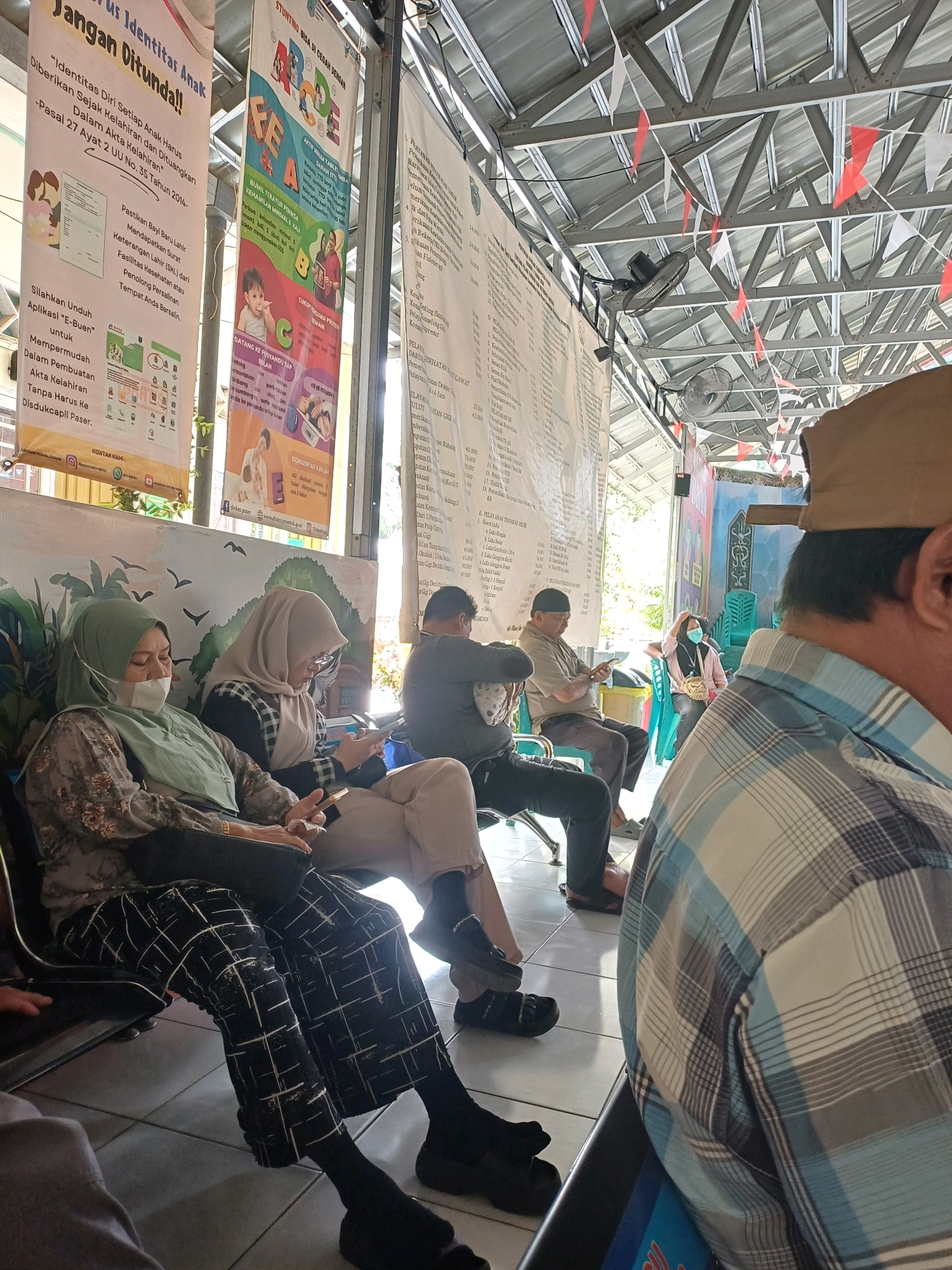 The image depicts a waiting area, possibly a public service office, in Pasir Belengkong, Indonesia. Several people are seated, appearing to wait for their turn. In the foreground, the back of a person wearing a blue and white checkered shirt is visible. To the left, two women are seated, both wearing hijabs. The woman on the far left is wearing a light green hijab and a patterned top, with a face mask covering her nose and mouth. She is looking down at her phone. Next to her, another woman in a beige hijab and a black and white patterned top is also looking at her phone.
In the background, more individuals are seated. A man in a grey shirt is looking at his phone, and another man in a light brown shirt is also seated, holding what appears to be a document. Further back, a woman in a pink top and a dark hijab is visible. The environment appears to be an open-air covered area with a metal roof structure. Red and white triangular flags are strung across the ceiling. Several posters or banners with text and images are hanging on the walls, some displaying Indonesian writing. The floor is tiled. Two electric fans are mounted on a structural beam. The overall impression is one of a government or community service office, likely a place for administrative tasks or consultations, given the presence of informational banners.