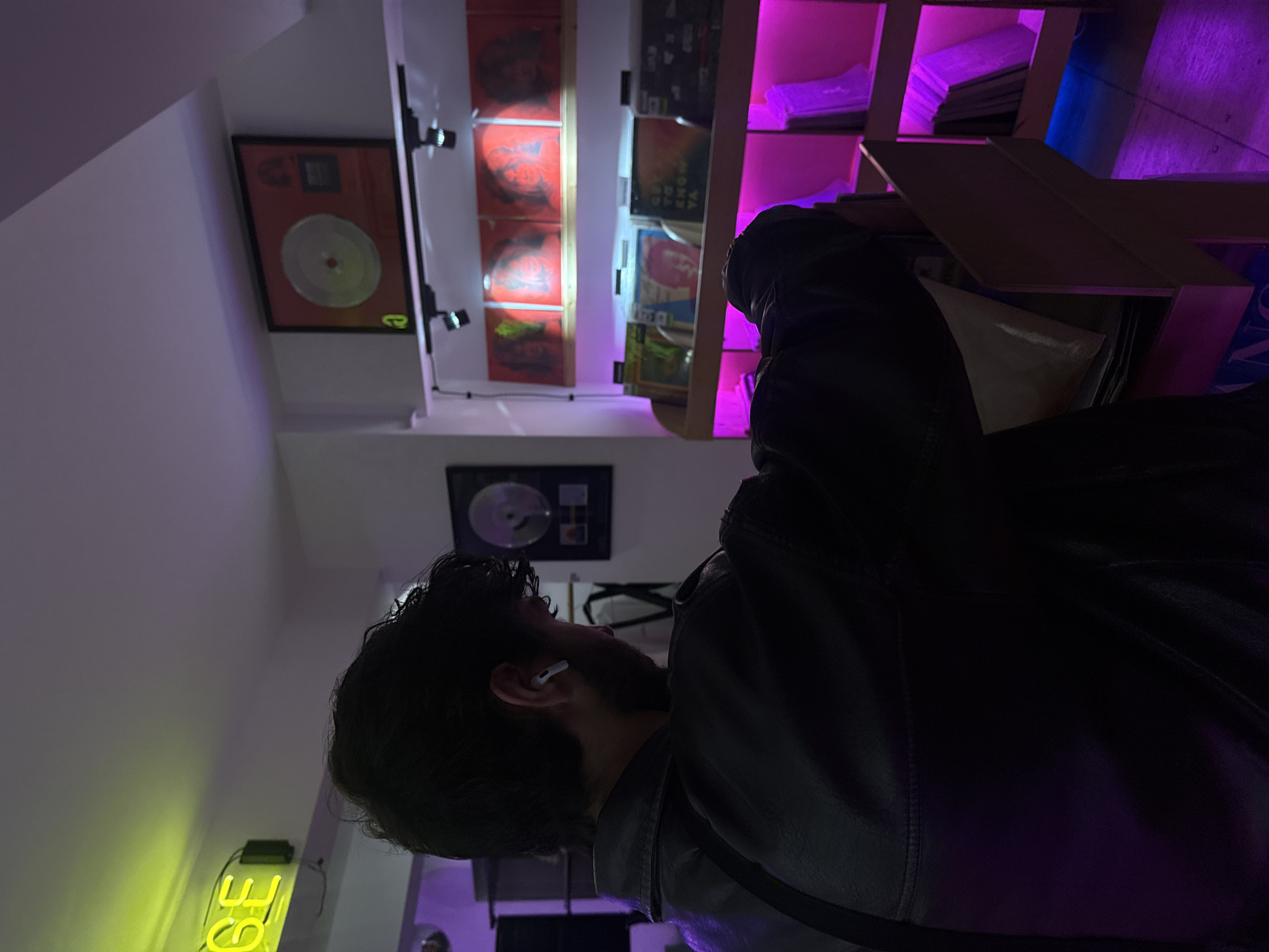 A low-angle shot reveals a man with dark hair and a beard, wearing an AirPod in his left ear and a dark jacket, standing inside a record store in Yerevan, Armenia. He is looking at a shelf of records illuminated with purple LED lights. On the wall behind the shelves are several framed records and a yellow neon sign is visible in the background.