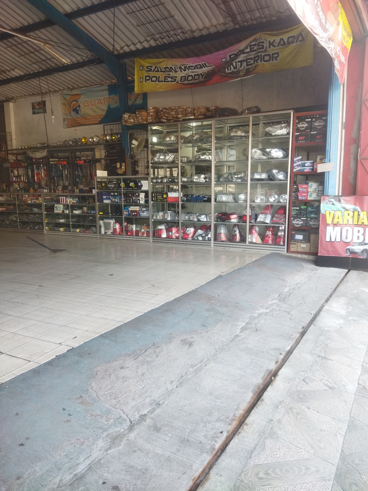 The image shows the interior of a car parts shop in Wonogiri, Indonesia. The shop is filled with shelves of car parts, including headlights, taillights, and other accessories. The shelves are made of glass and metal, and the parts are neatly displayed. There is a banner hanging on the wall that reads "SALON MOBIL POLES BODY POLES KACA INTERIOR". In the foreground, there is a concrete ramp leading into the shop. The shop appears to be well-lit and organized. There are no people visible in the image.