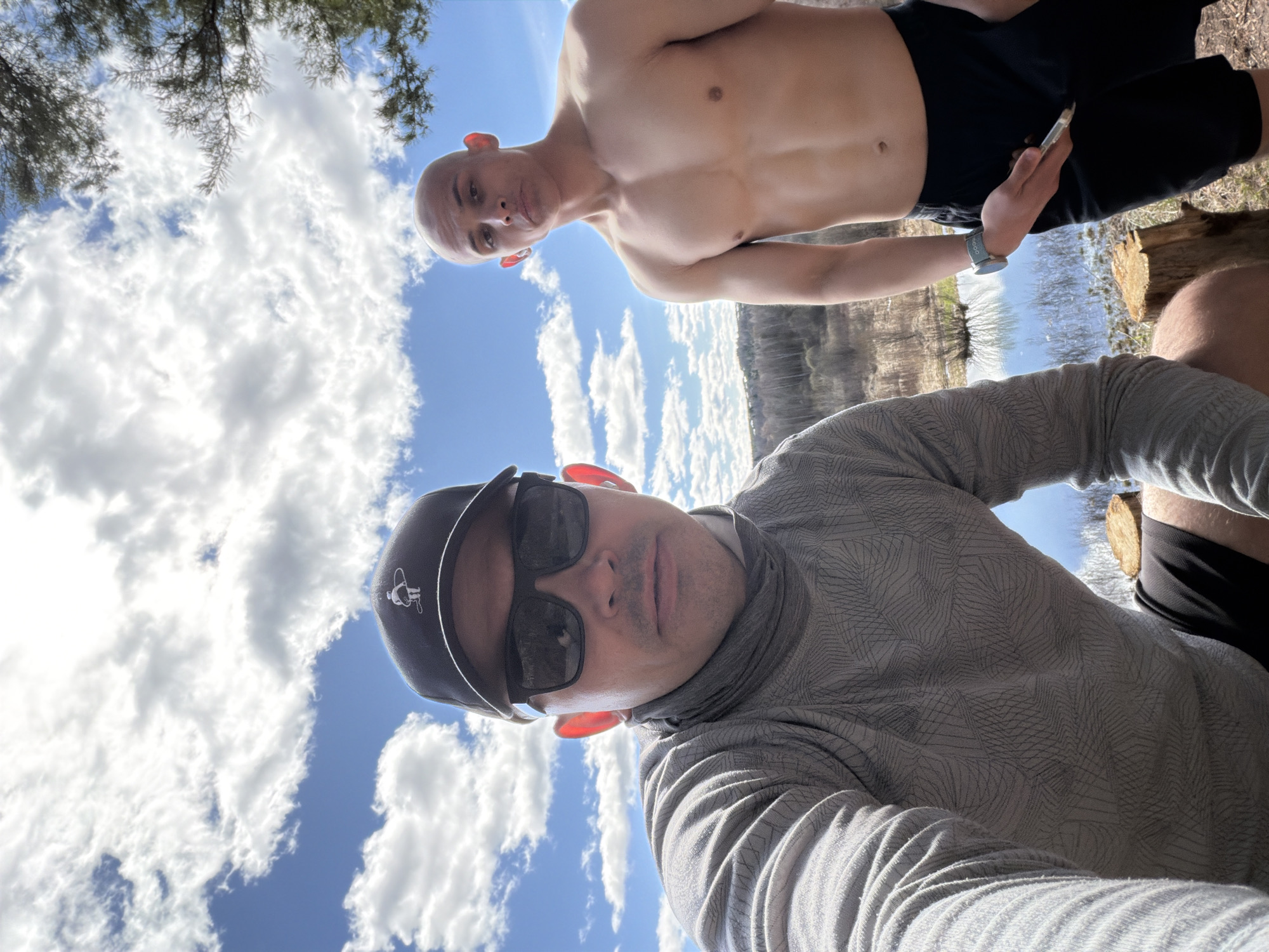 Two men are in the image, taken from a low angle. One man is shirtless and wears black shorts; he holds a small object in his right hand and wears a watch on his left wrist. The other man wears a gray patterned long-sleeved shirt, black shorts, a black cap with a logo, and sunglasses. The background shows a blue sky with white clouds, trees, and water. There is no information to discern the location of the image in Izhevsk, Russia.