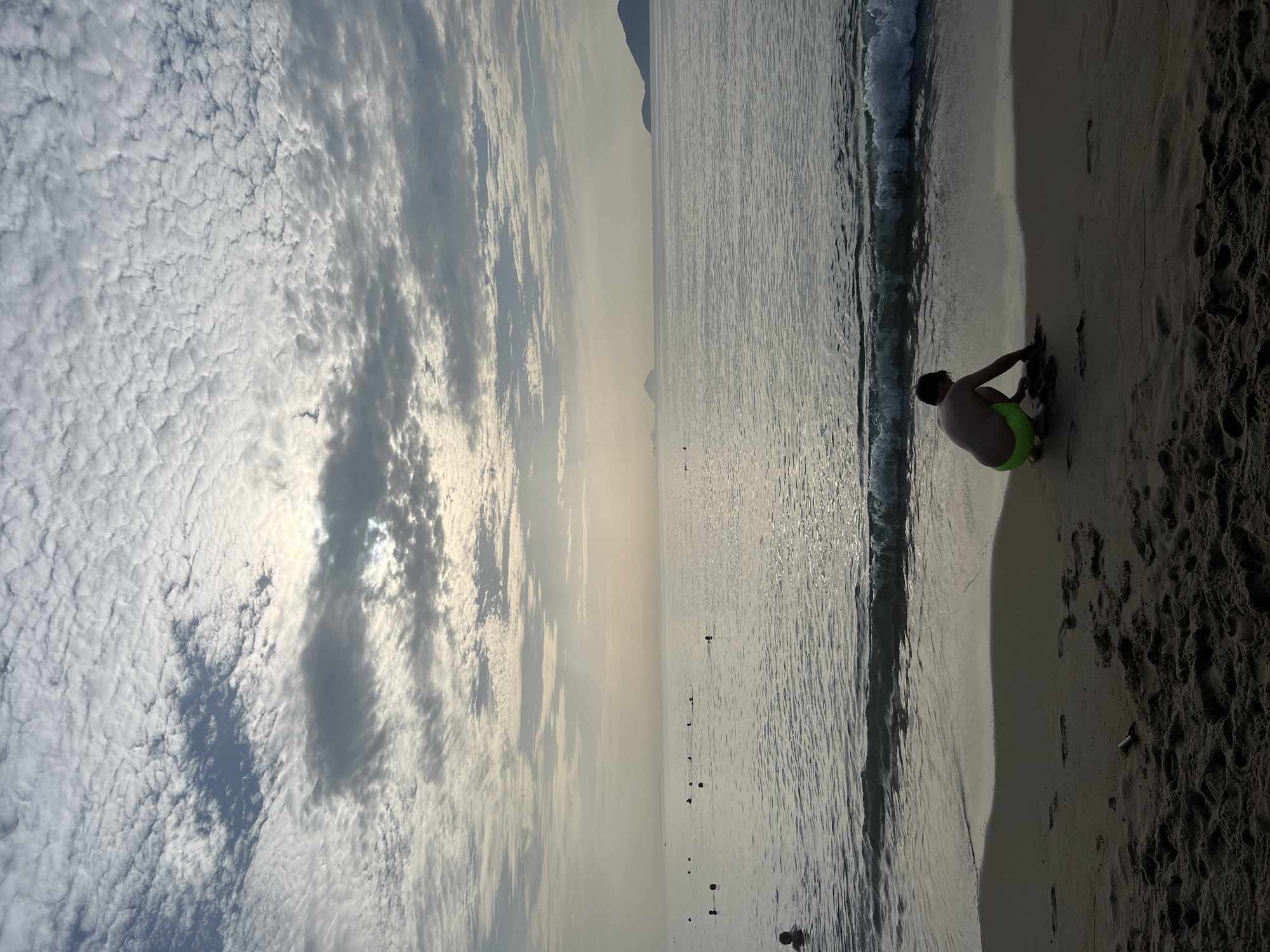 A lone young male, wearing bright green swim shorts, crouches on a sandy beach, appearing to play with the sand near the waterline. The tide is out, leaving wet sand exposed. Gentle waves lap at the shore. The ocean stretches out to the horizon, where distant islands are visible. The sky is filled with a dramatic pattern of white and gray clouds, with the sun partially obscured, creating a soft, diffused light. Several other people are scattered in the water, swimming or wading. The overall scene is peaceful and captures a moment of solitary activity on a beach. The location is inferred to be Nha Trang, Vietnam, given the visual characteristics of the beach and the presence of islands.