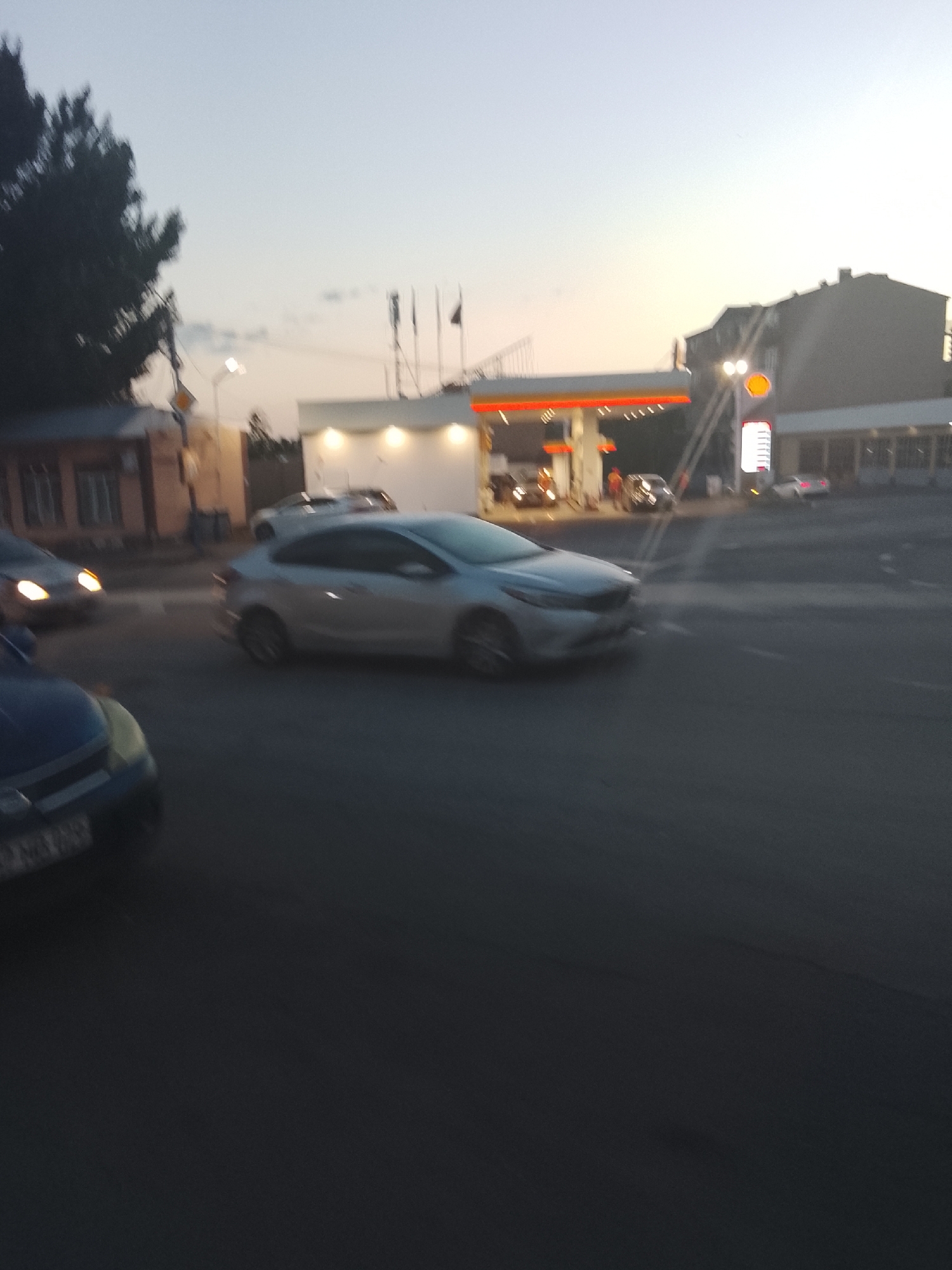 An outdoor, blurry, daytime scene depicts a gas station and surrounding street with several cars. In the foreground, a dark blue car is partially visible on the left, with its headlights on. To its right and slightly further back, a silver sedan is captured mid-turn, appearing to be a Kia Forte. Beyond the silver car, a white car with its headlights on is moving away from the camera. The gas station itself features a prominent red and yellow canopy with illuminated white stripes. A tall, white pylon sign with a Shell logo is visible to the right of the canopy, displaying vertical lines of pricing information. Two individuals in what appear to be uniforms are standing near a pump, and other vehicles are visible at the pumps. The background shows a brick building on the left and a larger, darker building on the right. The sky is a pale blue with hints of the setting sun. The scene is set on an asphalt road.