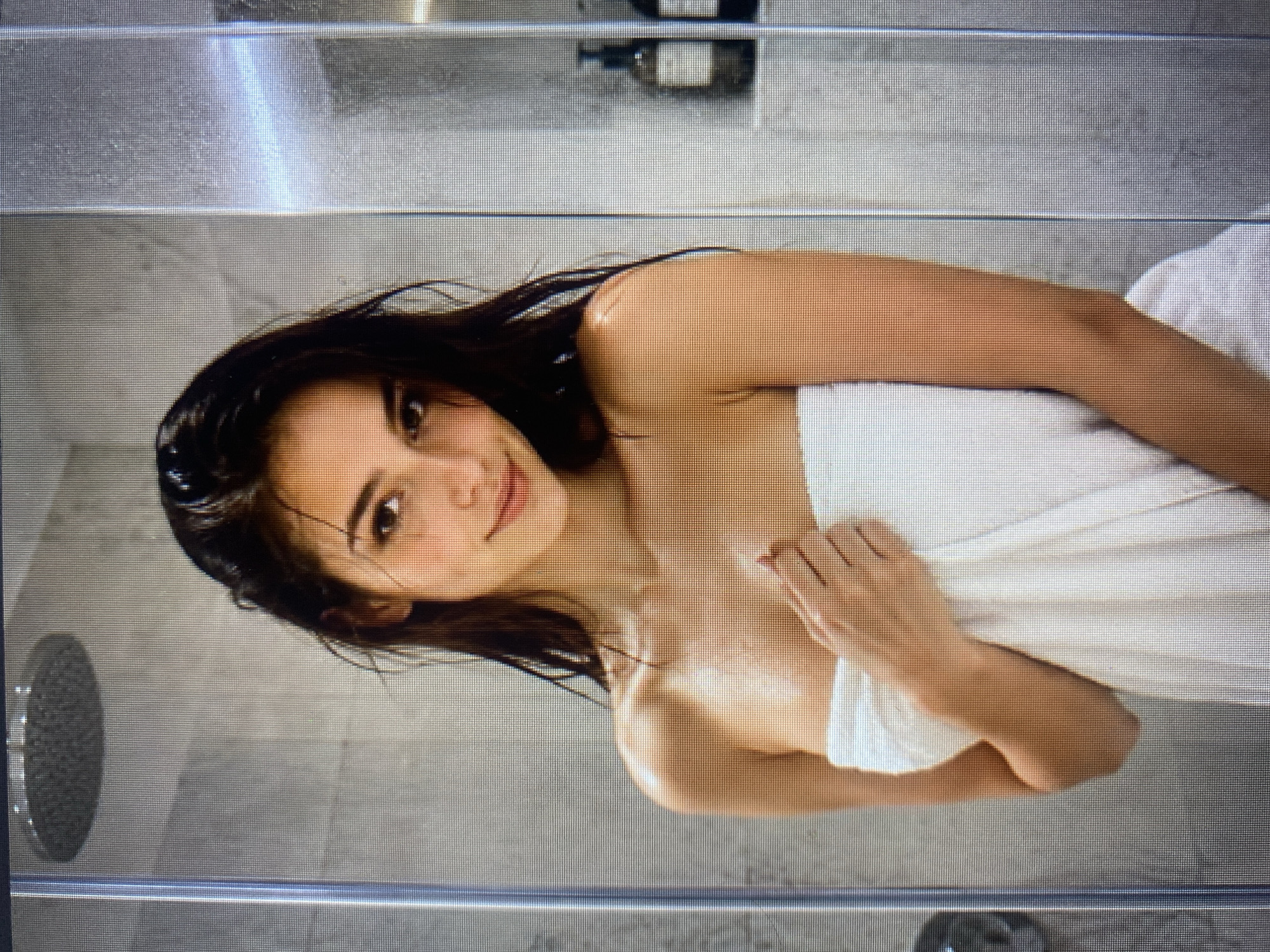 A woman stands in a shower, wrapped in a white towel. She has dark, wet hair and is looking at the camera with a slight smile. Her skin is glistening, suggesting she has just showered. The shower area features a modern showerhead and tiled walls. To the right, on a shelf, are two dark bottles, likely shampoo or soap. The lighting is soft and diffused.