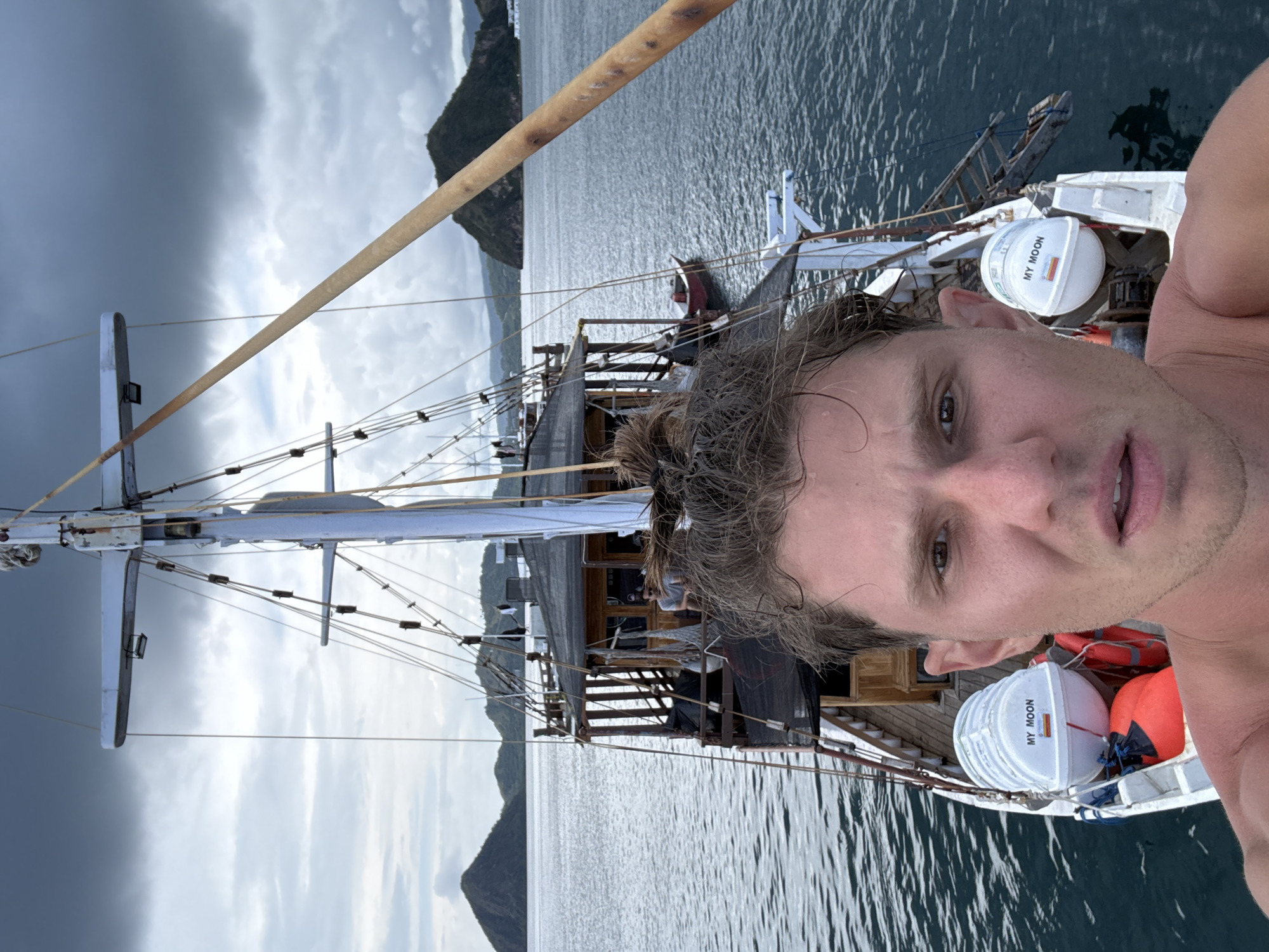 A disoriented, selfie-style photograph shows a shirtless Caucasian male with wet hair pulled into a bun. He is positioned on the bow of a boat, with the foreground dominated by his face and shoulder. The boat is named "My Moon", as labeled on one of the life rafts. The boat has a tall mast with white supports and rigging.
The background features water, mountains, and a cloudy sky. This appears to be the city of Pasir Putih, Indonesia, based on the geographical features of the surrounding landscape.