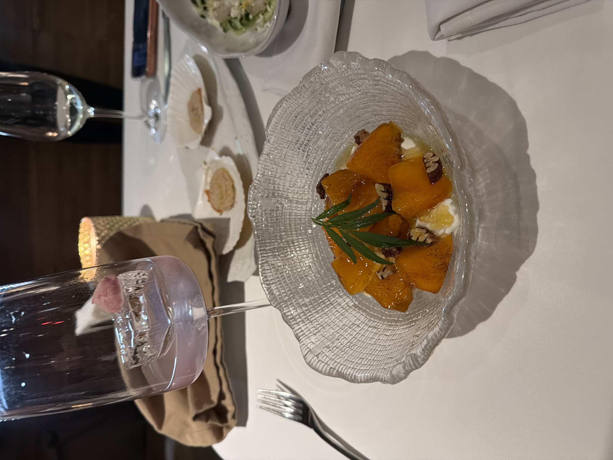 A restaurant table setting is depicted in this image. A glass bowl contains chunks of orange fruit, possibly persimmon, interspersed with dollops of white cream or cheese and topped with green leaves, possibly herbs. To the left of the fruit dish, there is a glass with a pink-colored ice cube and a fork. Further to the left, there is another glass of clear liquid. Two oyster shells, each containing a portion of food, are visible between the glasses. A salad bowl is partly visible in the upper left corner. A folded white napkin rests in the upper right corner of the scene. There are no people visible in the image. The location of the image is not specified.