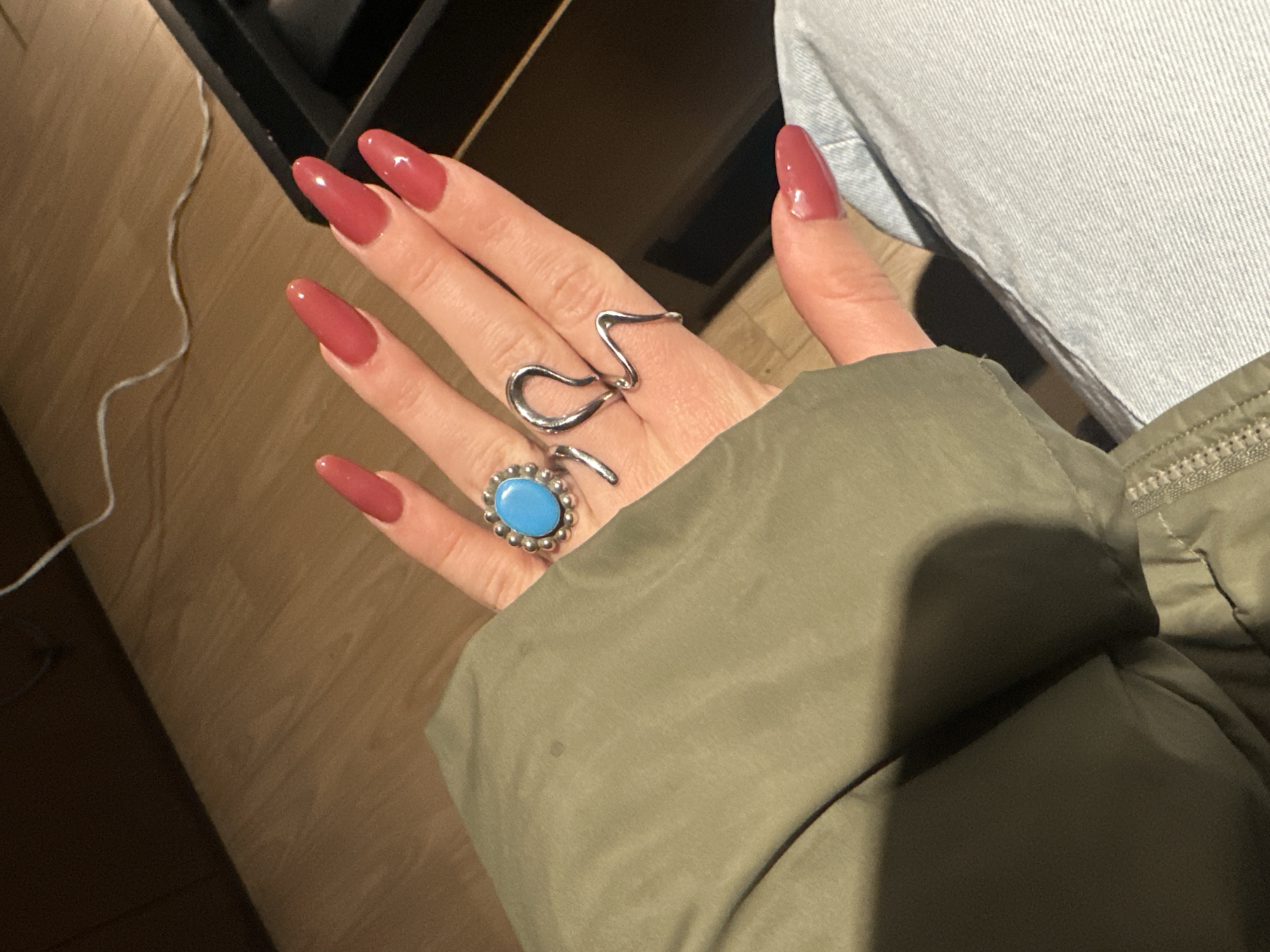 A hand with red manicured nails is adorned with two silver rings, one featuring a blue stone and the other a snake design. The hand is partially covered by a green jacket sleeve. The setting appears to be indoors with a wooden floor and the edge of a piece of furniture visible in the background. A portion of a blue garment, possibly denim jeans, can also be seen. The location is not discernible based solely on the visual information provided.
