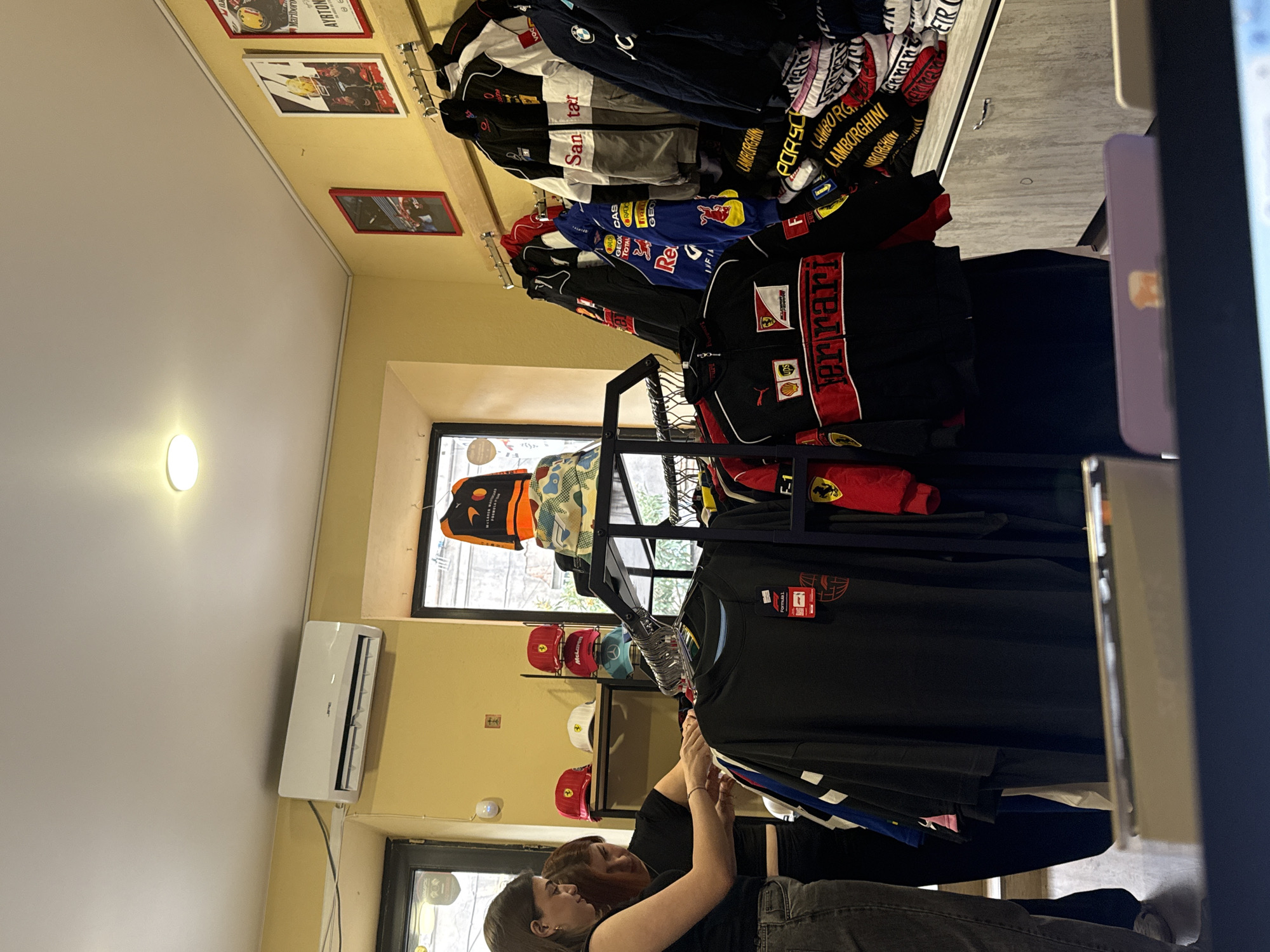 In an indoor retail environment with yellow walls and a white ceiling, two women are browsing clothing items. One woman, on the left, is wearing a black t-shirt and grey jeans. The other woman, in the center, is wearing a black t-shirt and black pants. They are standing in front of a rack of black and red motorsport-themed jackets and shirts, along with a display of caps. To the left, an air conditioning unit is mounted high on the wall. The background includes a window with a view of trees and a shelf displaying several caps, including red Ferrari-branded ones and a blue one. On the wall above the clothing rack, framed posters with racing imagery are displayed. The overall scene depicts a clothing store specializing in motorsport apparel.