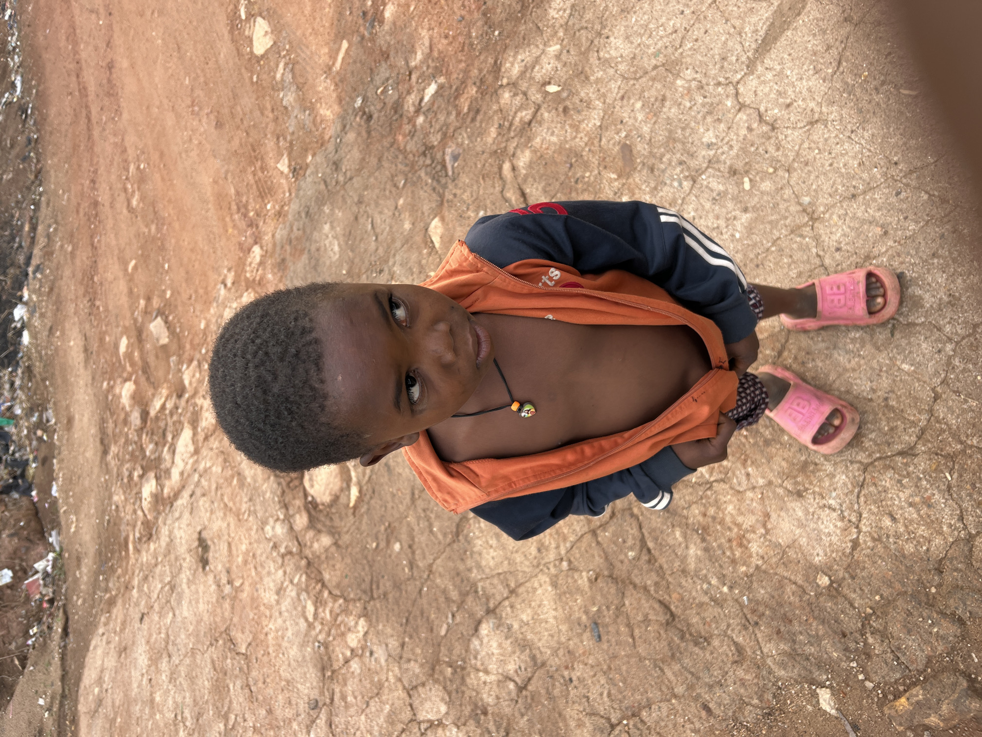 A young child, likely a boy, with dark skin and short, dark hair, stands looking up directly at the camera. The photo is taken from a high angle, capturing him from above. He is wearing an orange and dark blue jacket or hoodie, unzipped to reveal his bare chest. Around his neck, he has a black string necklace with a small, colorful bead or pendant. On his feet, he wears bright pink slide sandals, one of which appears to have a "BB" logo or similar text on the strap.
The child stands on a textured, reddish-brown ground, which appears to be a mix of dirt and cracked, weathered concrete or pavement. Small stones and debris are scattered across the surface. To the left side of the frame, there is a blurry area containing what looks like litter and disturbed ground, suggesting an unkempt or roadside environment. The lighting is bright and even, indicating it is daytime. The child's expression is neutral and calm, gazing upwards. The scene takes place in Gura Top, Nigeria.