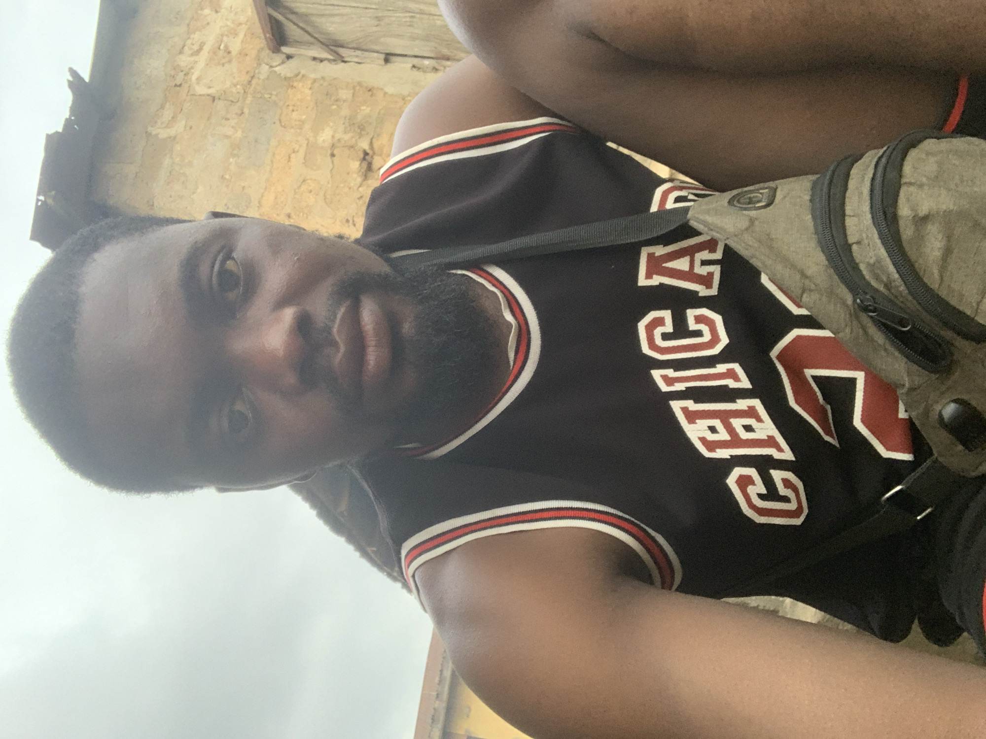 A man is pictured in a close-up, low-angle selfie. He is wearing a black sleeveless basketball jersey with "CHICAGO" written in red and white lettering across the chest, and a number "23" below it. A dark strap from a messenger bag crosses his chest, and the bag itself, a muted green color with zippers, is visible resting on his right leg. The man has dark skin, short black hair, and a beard. In the background, a textured, light-colored brick wall with some debris on top is visible, suggesting an outdoor urban setting. The sky is overcast and pale. This scene is located in Shagamu, Nigeria.