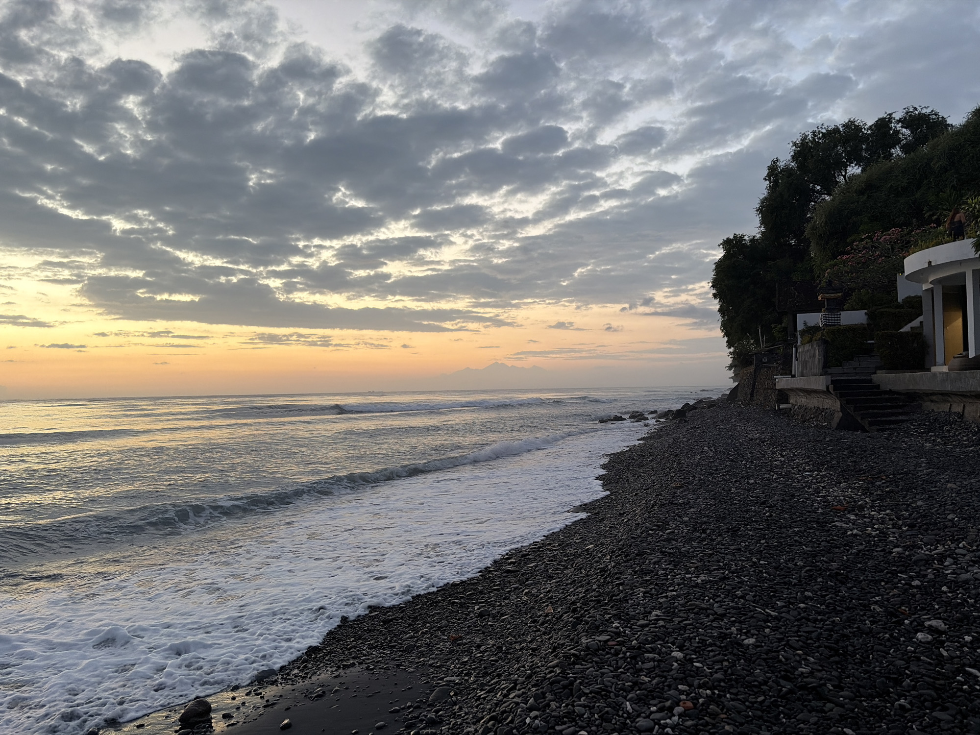 A view of a rocky beach at dusk in Abang, Indonesia. The sky is filled with dramatic clouds, with a warm orange glow near the horizon. The ocean is calm, with gentle waves lapping at the shore. The beach itself is covered in dark, smooth pebbles. In the background, a lush green hillside is dotted with trees, and a modern building with white circular columns and a staircase leading up to it is visible. A lone figure, appearing to be a man in dark shorts, stands on a balcony of the building. The overall impression is one of natural beauty and serenity.