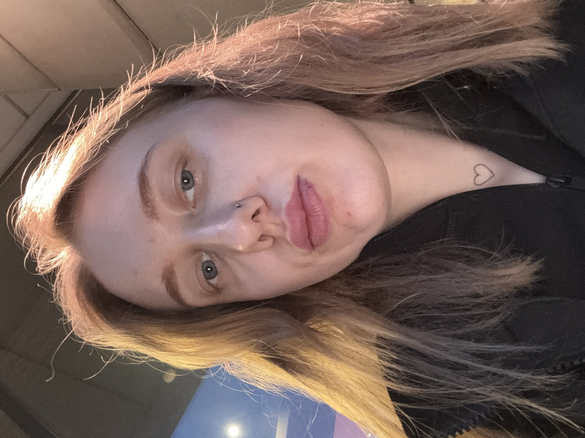 A selfie of a fair-skinned person with light brown hair. The person has blue eyes, a piercing on the left side of their nose, and a small heart tattoo on the neck. The person is wearing a black jacket. The location appears to be indoors, with a blurry blue and pink surface visible in the lower part of the frame. No other people or objects are identifiable in the image. There is no indication this image was taken in Ol'ginskaya, Russia.