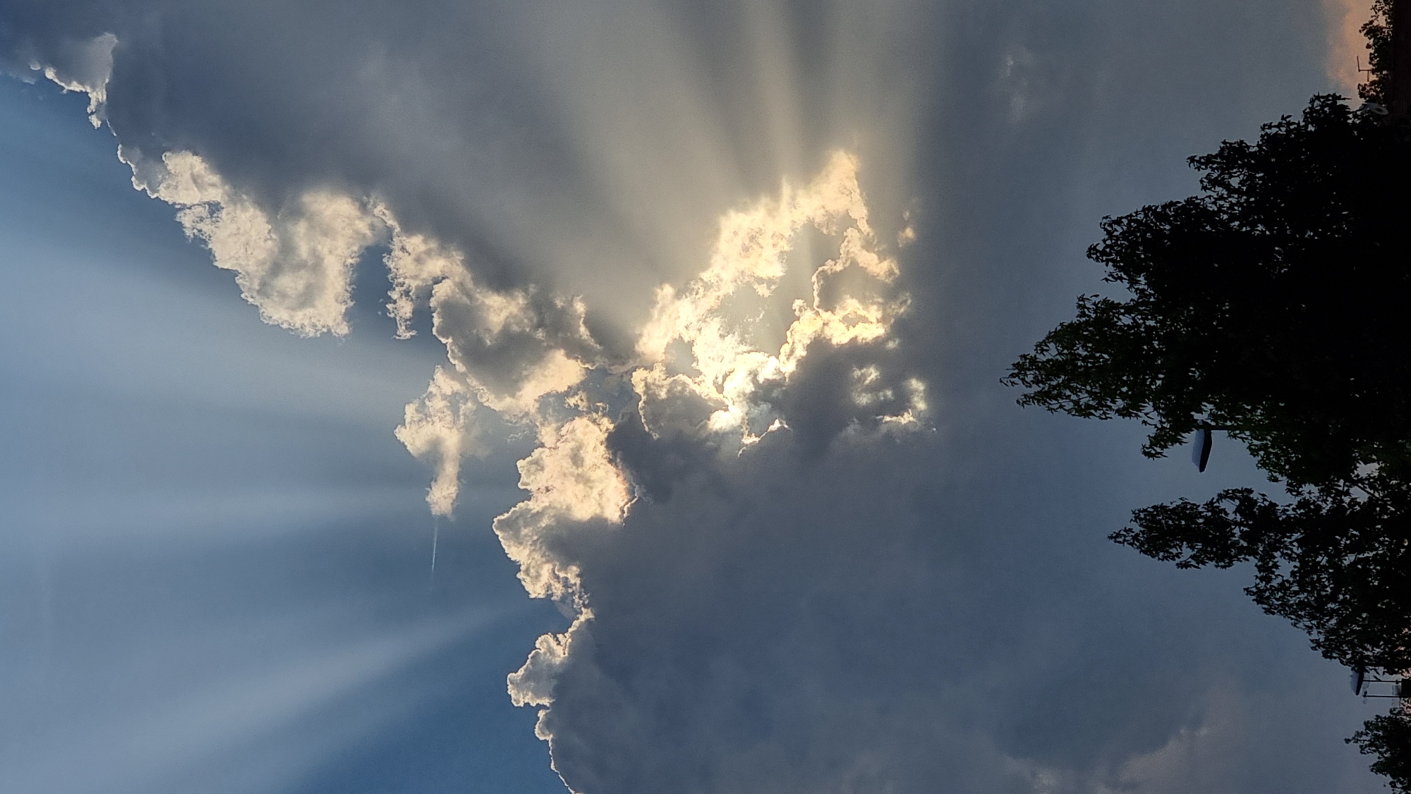 A low-angle, full-shot photograph captures a dramatic sky with crepuscular rays fanning out from behind a cluster of clouds. The sun is partially obscured, with bright light emanating from behind the dense cloud formations, creating a halo effect. The clouds are a mix of dark, heavy masses and thinner, illuminated edges. In the foreground, the dark silhouettes of trees and streetlights are visible against the twilight sky. The scene suggests an urban or suburban environment in Sant Joan Les Fonts, Spain, given the presence of streetlights.