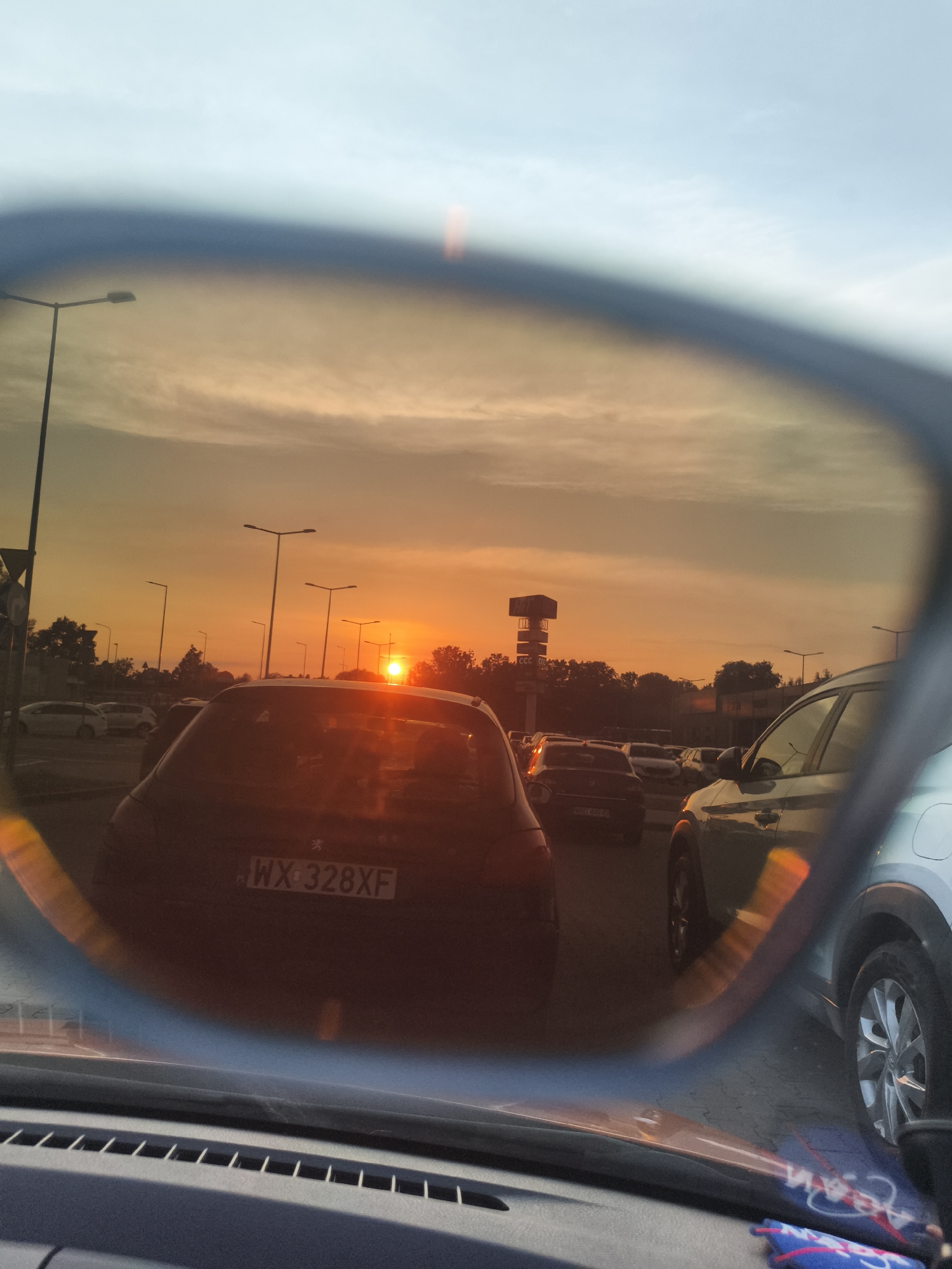 The image depicts a sunset viewed through a car's side mirror. The sky is filled with warm orange and yellow hues, indicating the sun is low on the horizon. Several cars are visible in the traffic jam, their shapes distorted by the mirror's curvature. Streetlights stand tall along the road, their lights illuminated against the dusky sky. In the background, a tall structure with "CCC" signage is visible, suggesting a commercial area. The scene appears to be on a road in Nowy Dwór Mazowiecki, Poland, possibly near a shopping center given the CCC branding. The overall impression is one of a typical evening commute amidst a scenic sunset.