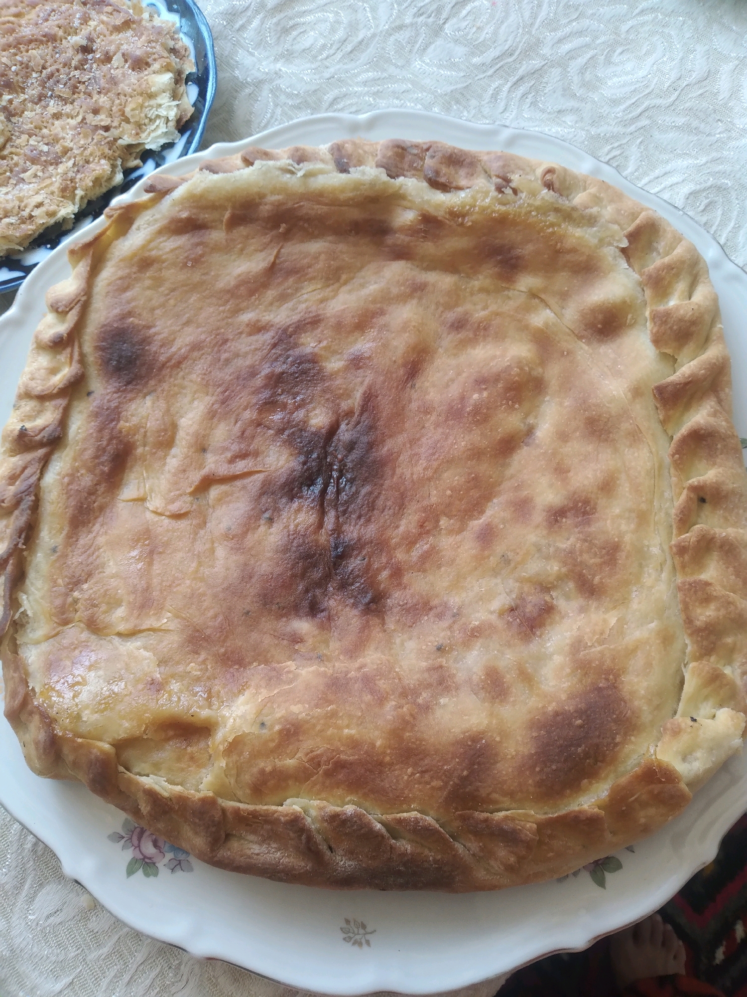 A rustic, golden-brown pie sits centrally on a white, scalloped plate decorated with faint floral patterns. The pie has a thick, braided crust and a slightly browned, textured filling visible through a few cracks. To the left, partially visible on a dark blue and white patterned dish, is a portion of another pastry, appearing flaky and topped with a light dusting. The scene is set on a textured, cream-colored tablecloth, with a sliver of a dark, patterned rug visible at the bottom right. The lighting suggests an indoor setting, possibly during the day. The overall impression is of a home-cooked meal.