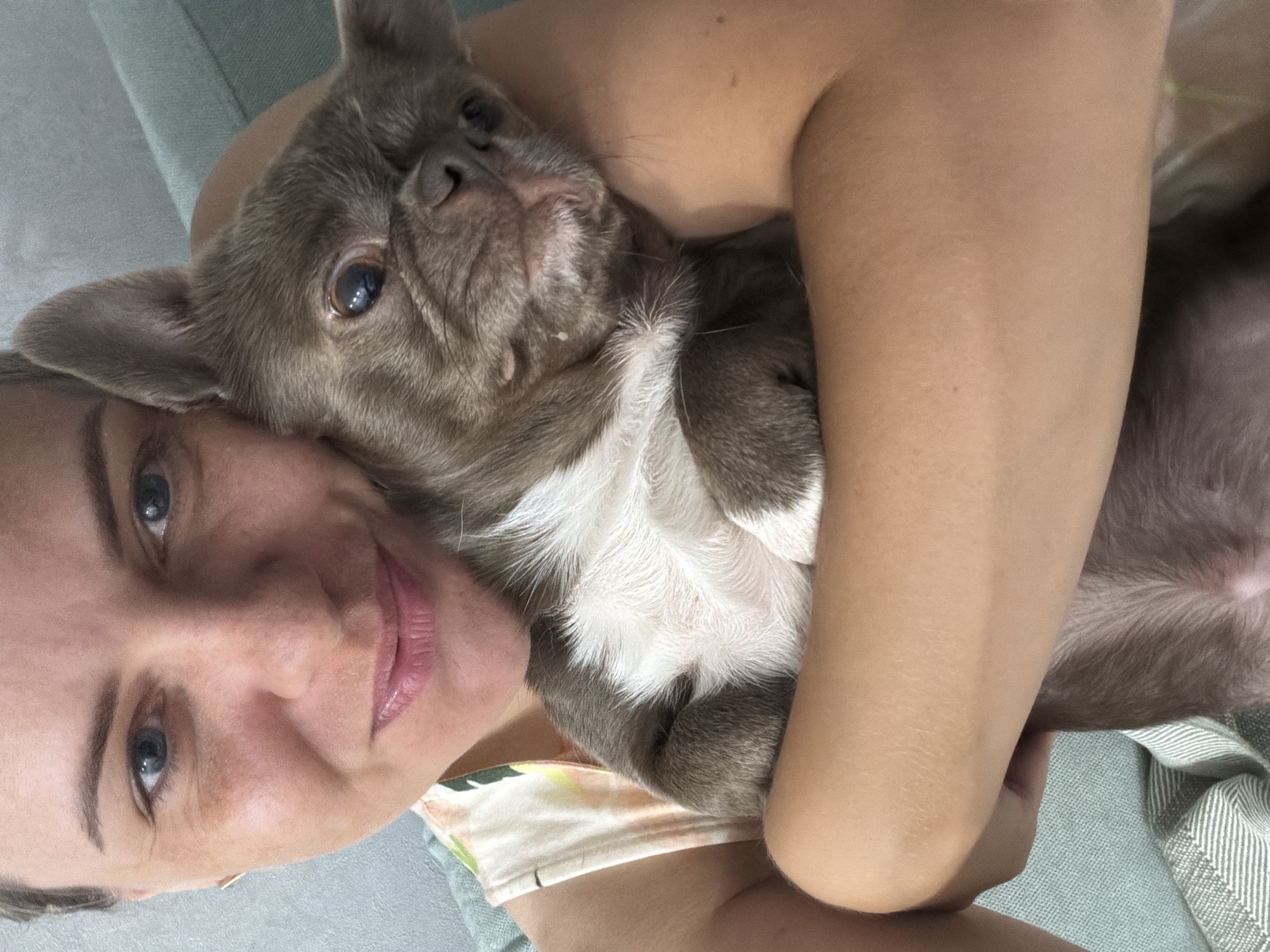 A woman is holding a French Bulldog. The woman has blonde hair pulled back and is looking at the camera with a slight smile. She is wearing a sleeveless top with a floral print. The dog is brown and white with large ears and is being held close to the woman's chest. The background appears to be a room with a light gray wall and a teal sofa. The image quality is not sharp enough to identify specific landmarks or details within the city of Votkinsk, Russia.