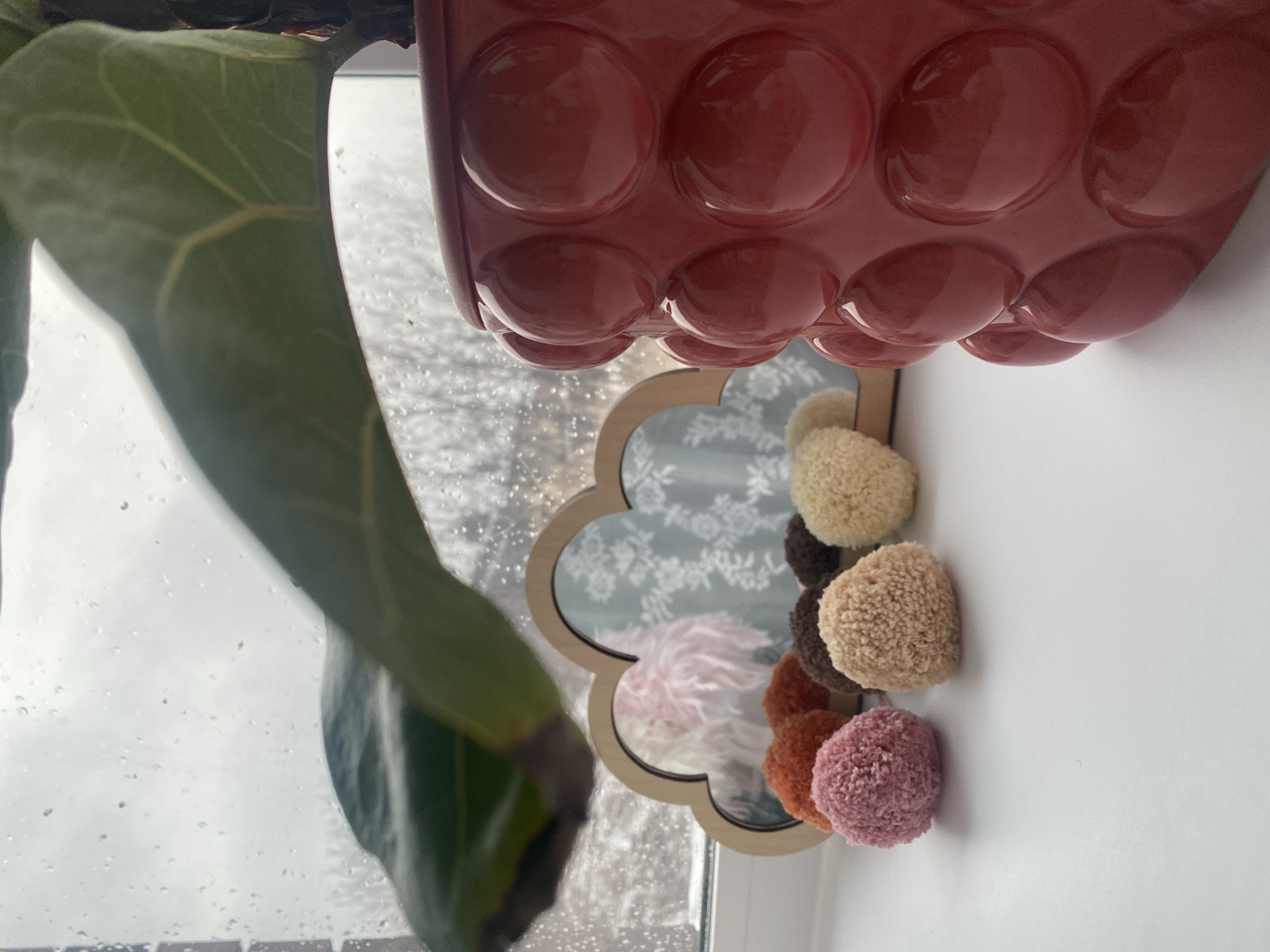 A sideways close-up shot shows a scene indoors, likely near a window. A large green plant leaf is visible on the left, partially obscuring the view. Above, a red, bubbly plant pot hangs down. In the center, a decorative mirror with pom-poms attached to its frame is present. The mirror reflects a window with raindrops and a view of what appears to be a building or scenery outside. The location, Izhevsk, Russia, is inferred from the context and metadata. The scene lacks human subjects or specific actions.