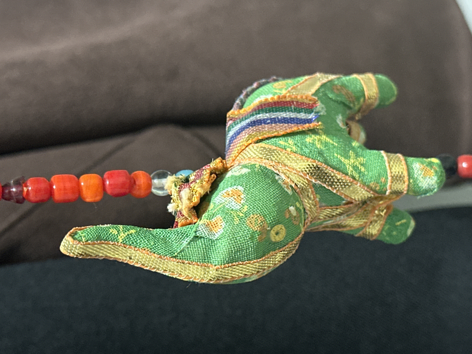 The image shows a close-up of a green fabric elephant figurine, adorned with gold trim and a colorful striped saddle. The figurine is hanging, attached to a beaded string. The beads are red, orange, and clear. The background consists of a dark gray fabric on the left and a textured brown fabric on the right. There is no identifiable information to suggest a specific location in Nevinnomyssk, Russia.