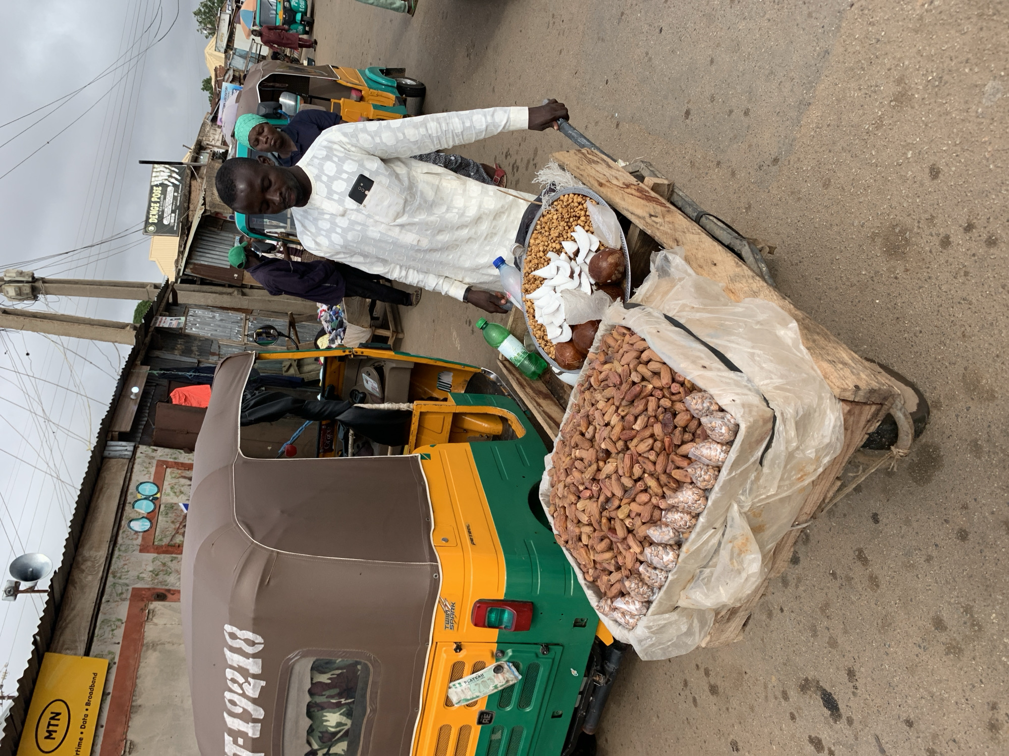 The image captures a vibrant street scene in Gura Top, Nigeria, centered around a male vendor selling goods from a wooden pushcart. The vendor, wearing a white patterned long-sleeved shirt and dark trousers, stands behind his cart, looking towards the camera.
His wooden cart is laden with various items: a large silver tray holds roasted chickpeas or groundnuts alongside fresh coconut slices and whole coconuts. Next to this, a voluminous transparent plastic bag is filled with numerous dates, with smaller, individually packaged portions also visible within. Two plastic bottles, one blue and one green, are placed among the goods.
To the vendor's left, a green and yellow "Keke Napep" (motor-rickshaw) is parked, featuring the text "F-19218," "TOWNS SPARK," "PETROL," and "PLATEAU" on its body. The street surface is an unpaved or worn asphalt road. The background reveals a lively street lined with simple, single-story buildings, some with corrugated iron roofs, indicative of a local market setting. A concrete utility pole with electrical wires stands prominently on the left.
Visible textual cues include a "DENGE POSE" signboard on a building and a yellow "MTN Airtime Data Broadband" sign attached to a pole. The scene unfolds under an overcast sky, suggesting a cloudy daytime. Several other people are visible in the background, contributing to the bustling atmosphere of the market.